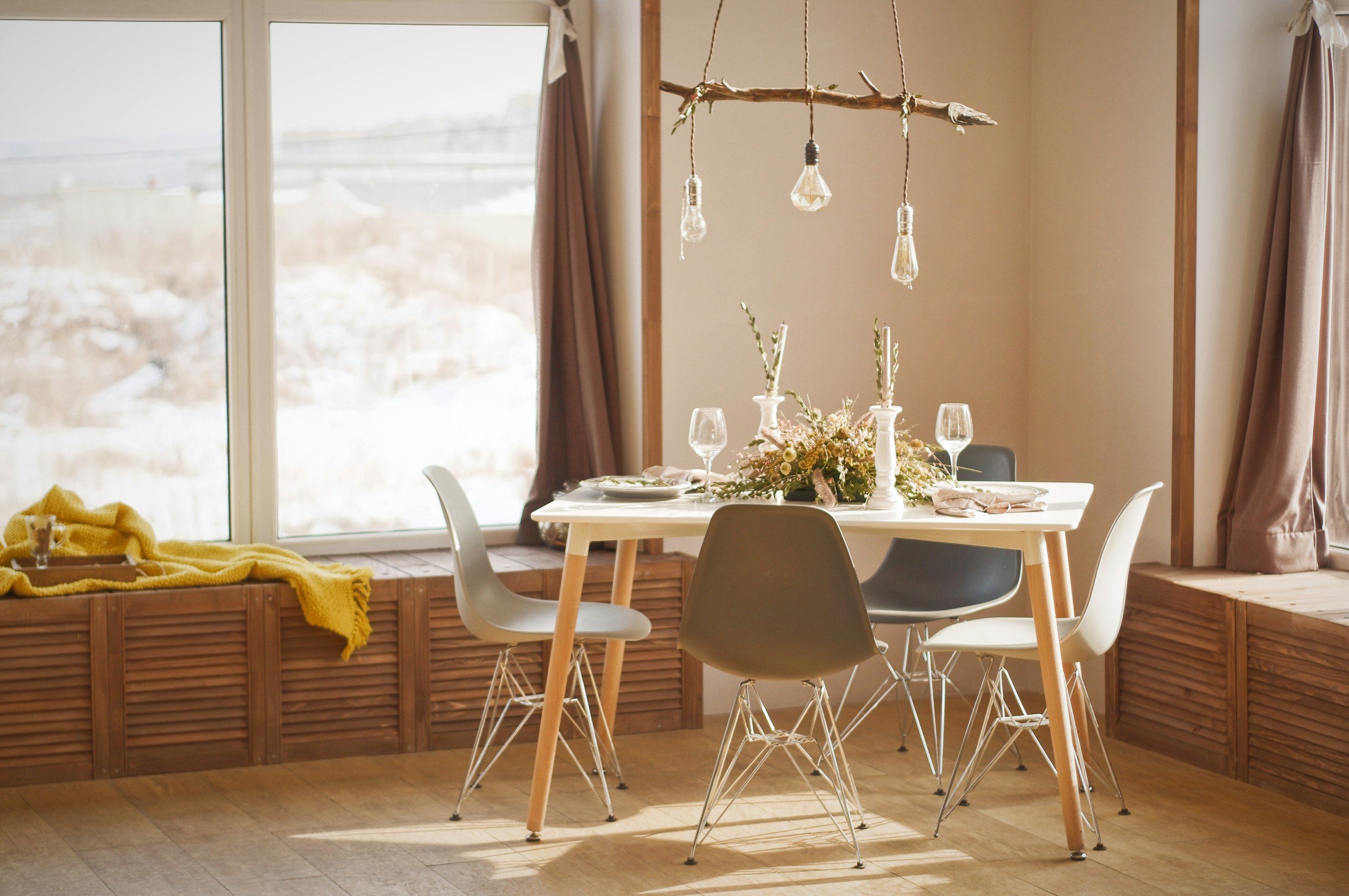 A dining area with a white table decorated with a floral centerpiece, surrounded by modern chairs. A branch with hanging light bulbs is suspended above the table. Large windows allow natural light, and pink curtains frame the windows. A yellow blanket is draped over a ledge.
