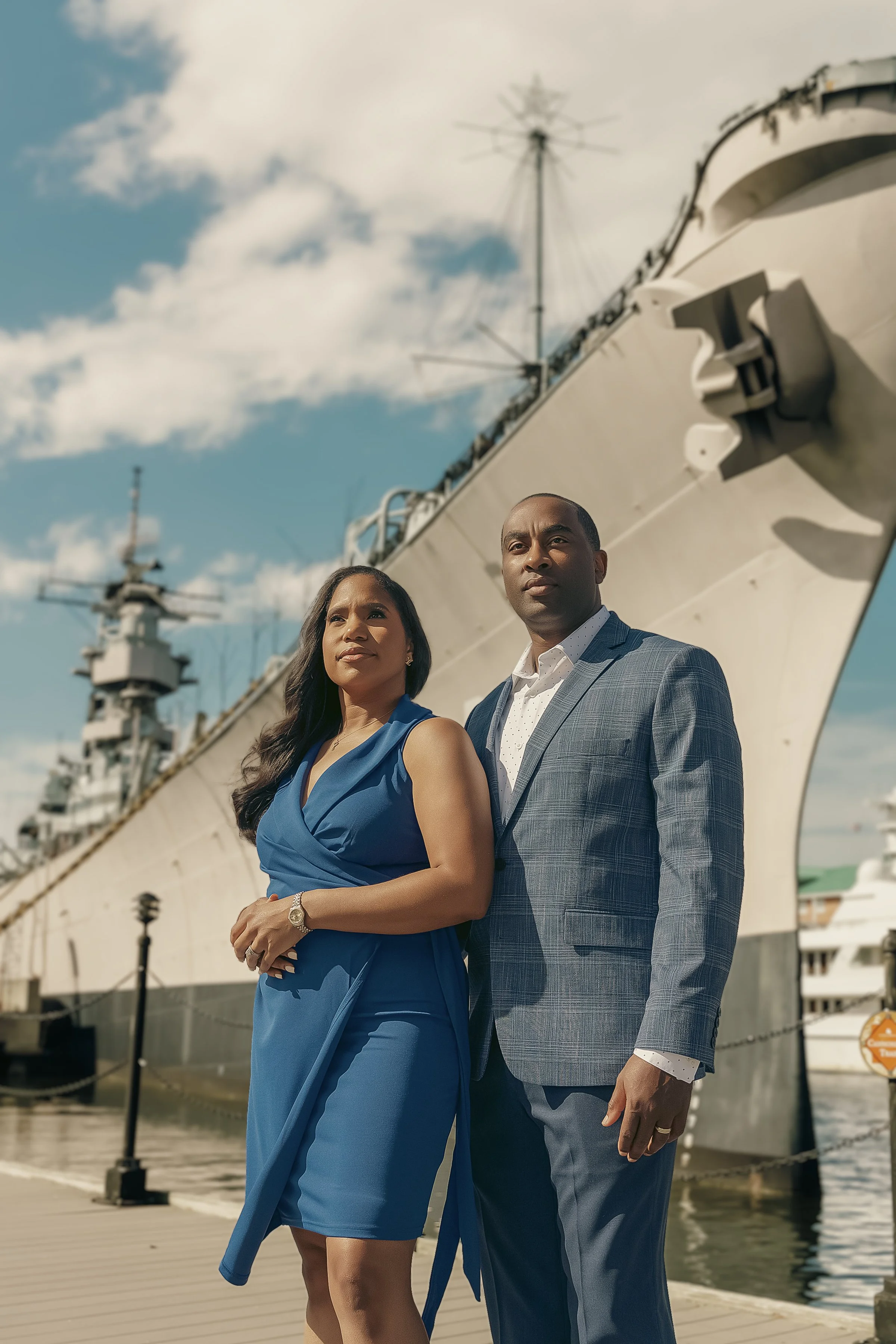 A man and woman dressed in formal attire standing on a dock near a large ship with a white hull, against a blue sky with scattered clouds.