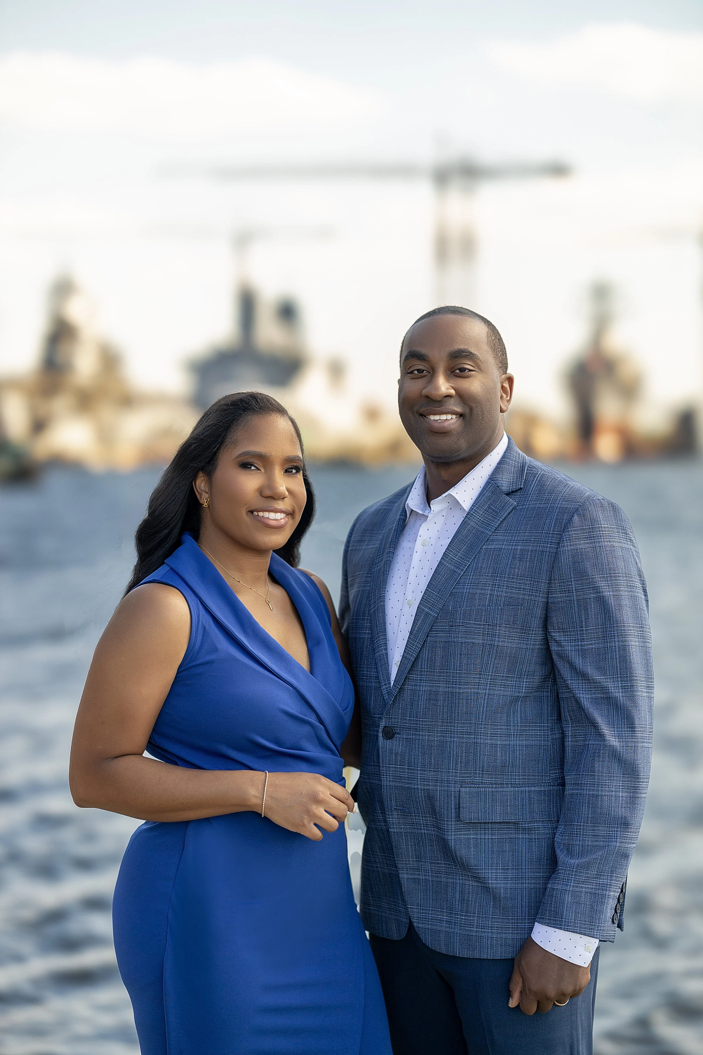 A well-dressed African American man and woman standing close together outdoors near water with ships and cranes in the background, smiling at the camera.