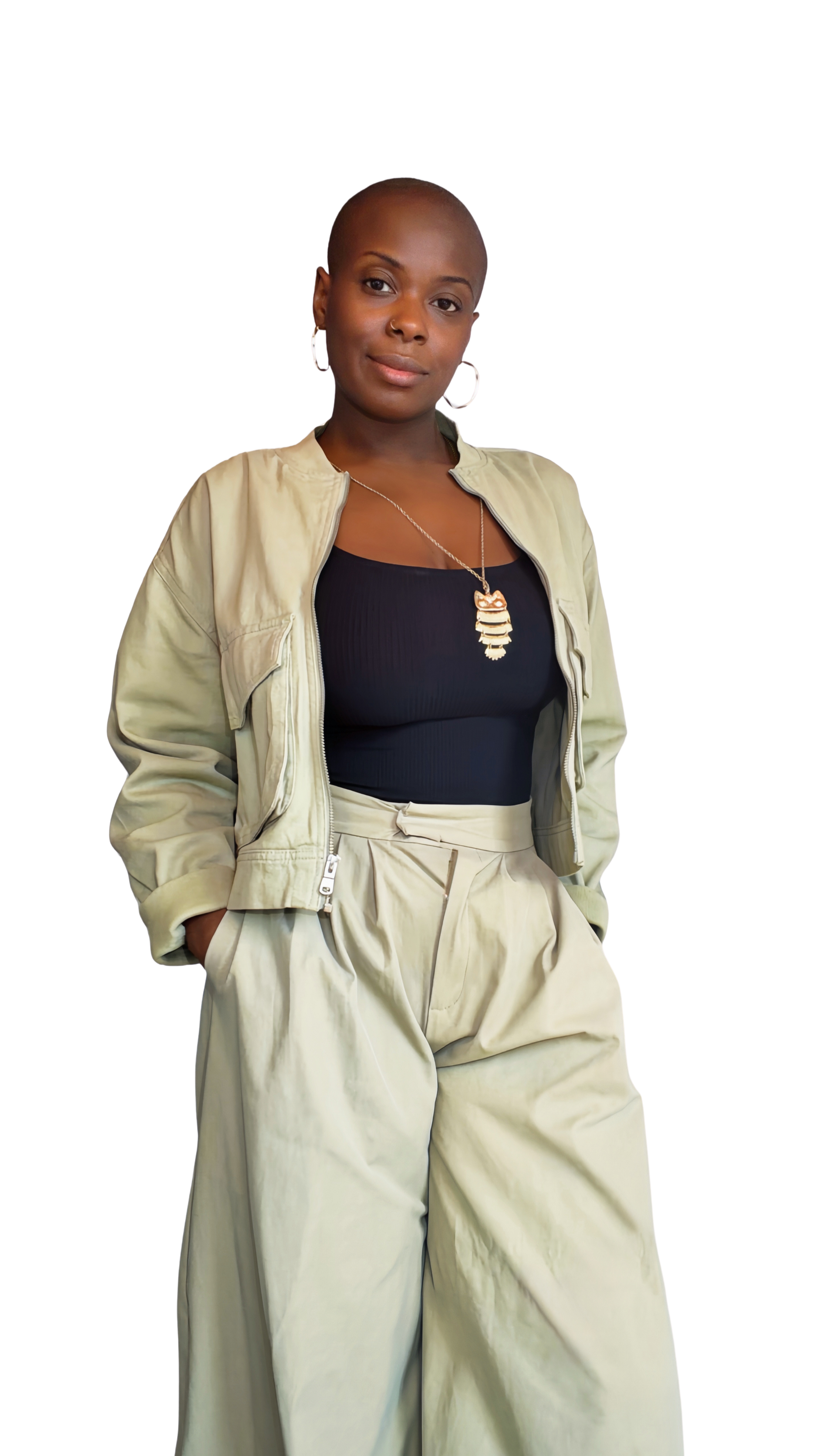 Une femme, portant une veste beige, un haut noir, et un pantalon ample beige, pose avec confiance. Elle a la tête rasée, porte des boucles d'oreilles, un collier avec un pendentif orné, et affiche un regard sûr.