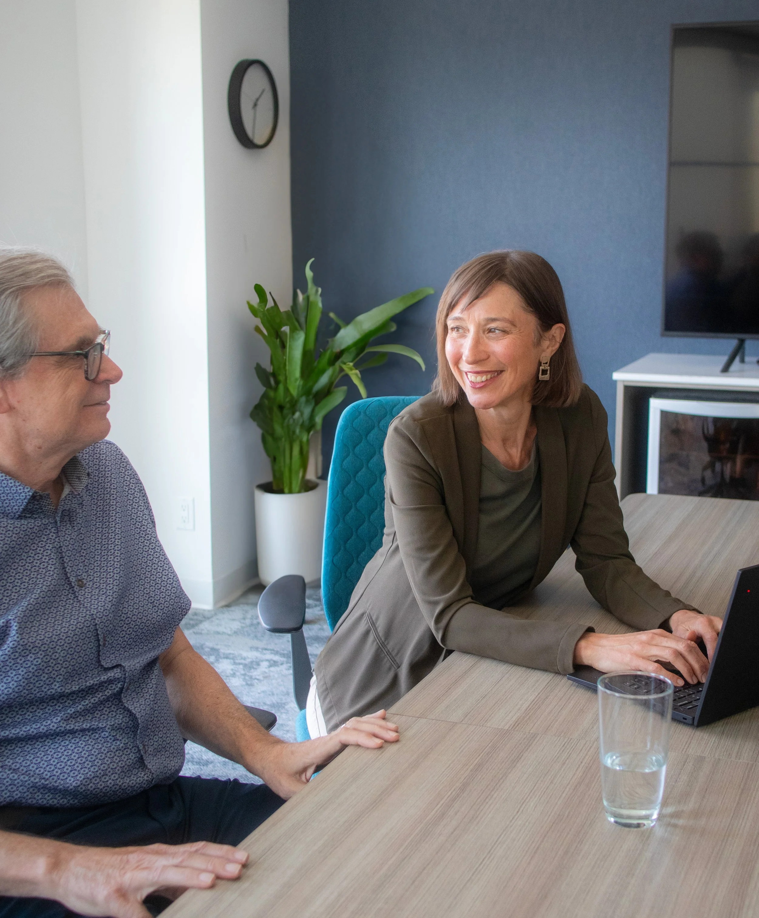 Two people sitting at a conference table, one with gray hair and glasses, the other smiling at a laptop, with a potted plant, a glass of water, and a TV on a stand in the background.