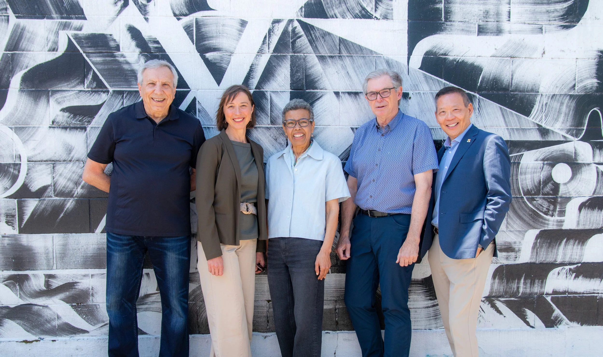 Five diverse adults stand together in front of a black-and-white abstract mural, smiling at the camera.