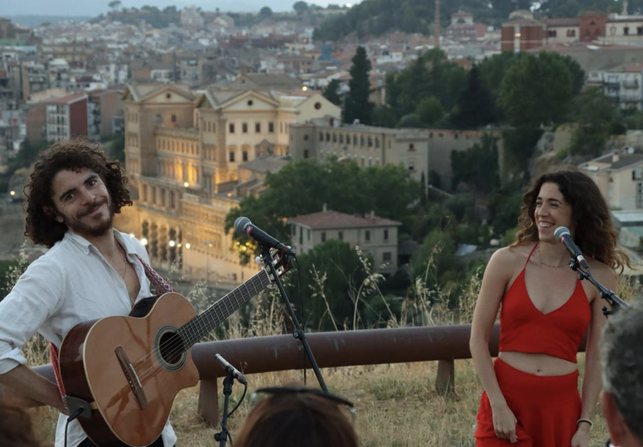 Claudia Bloom y Sean Hauser sonriendo entre guitarras y micrófonos durante una actuación musical al aire libre.