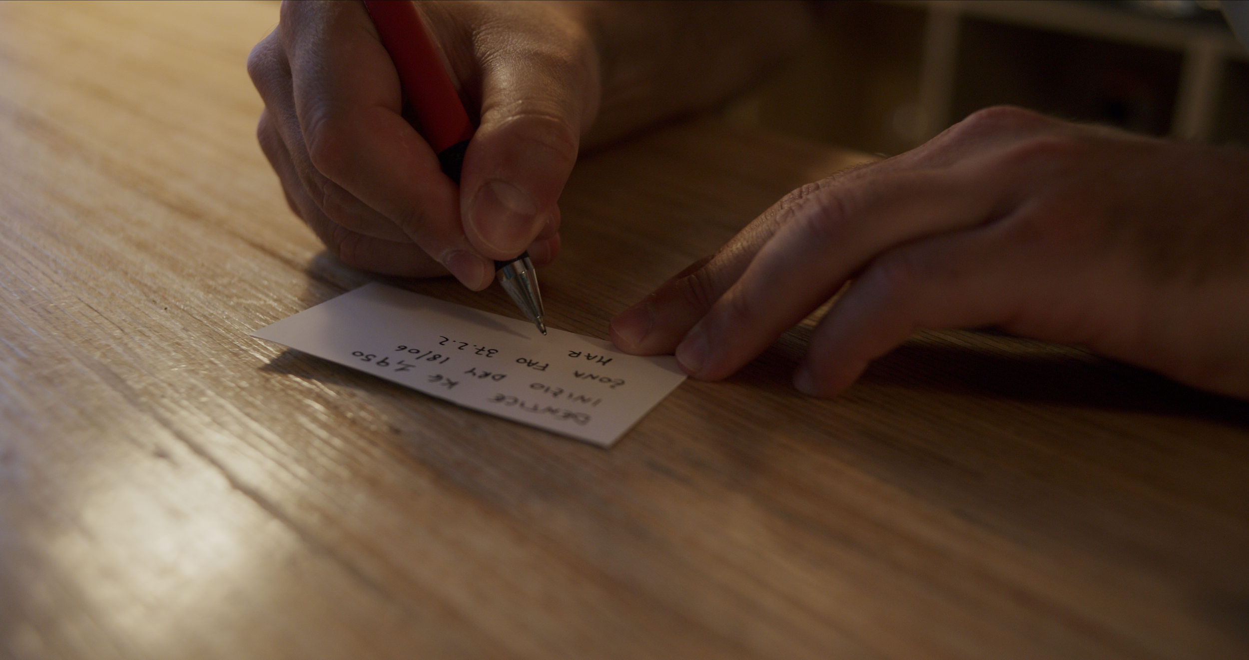 A person writing on a small piece of paper with a pen, on a wooden surface.