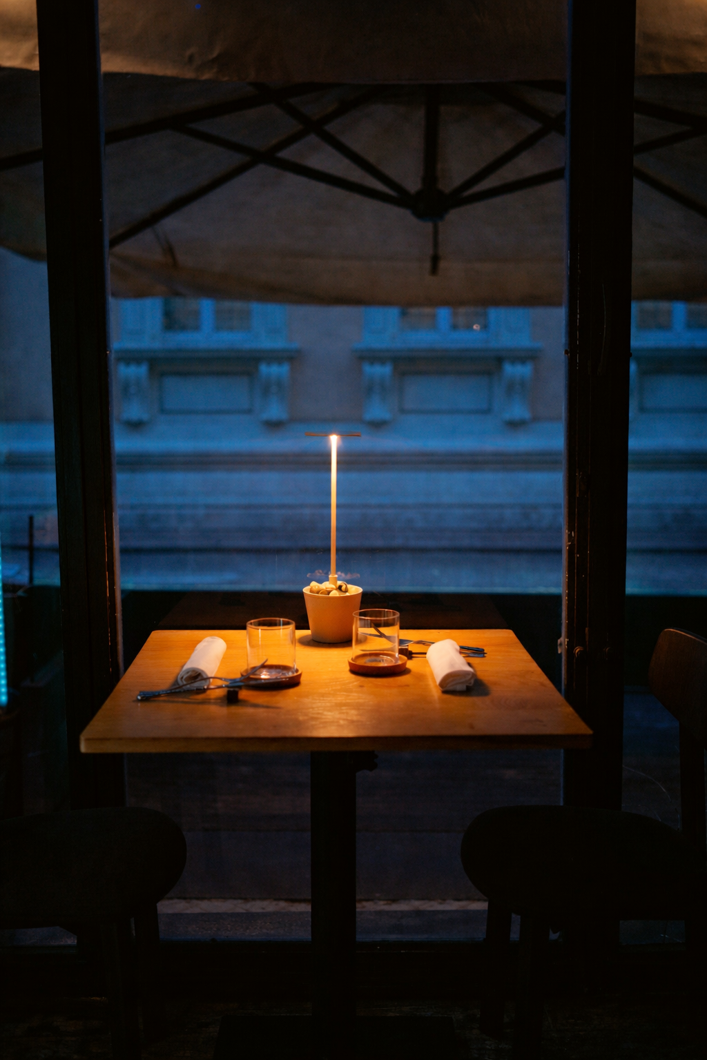A restaurant table set with two glasses, two napkins with cutlery, and a potted plant with a candle on top, under a large outdoor umbrella at dusk.