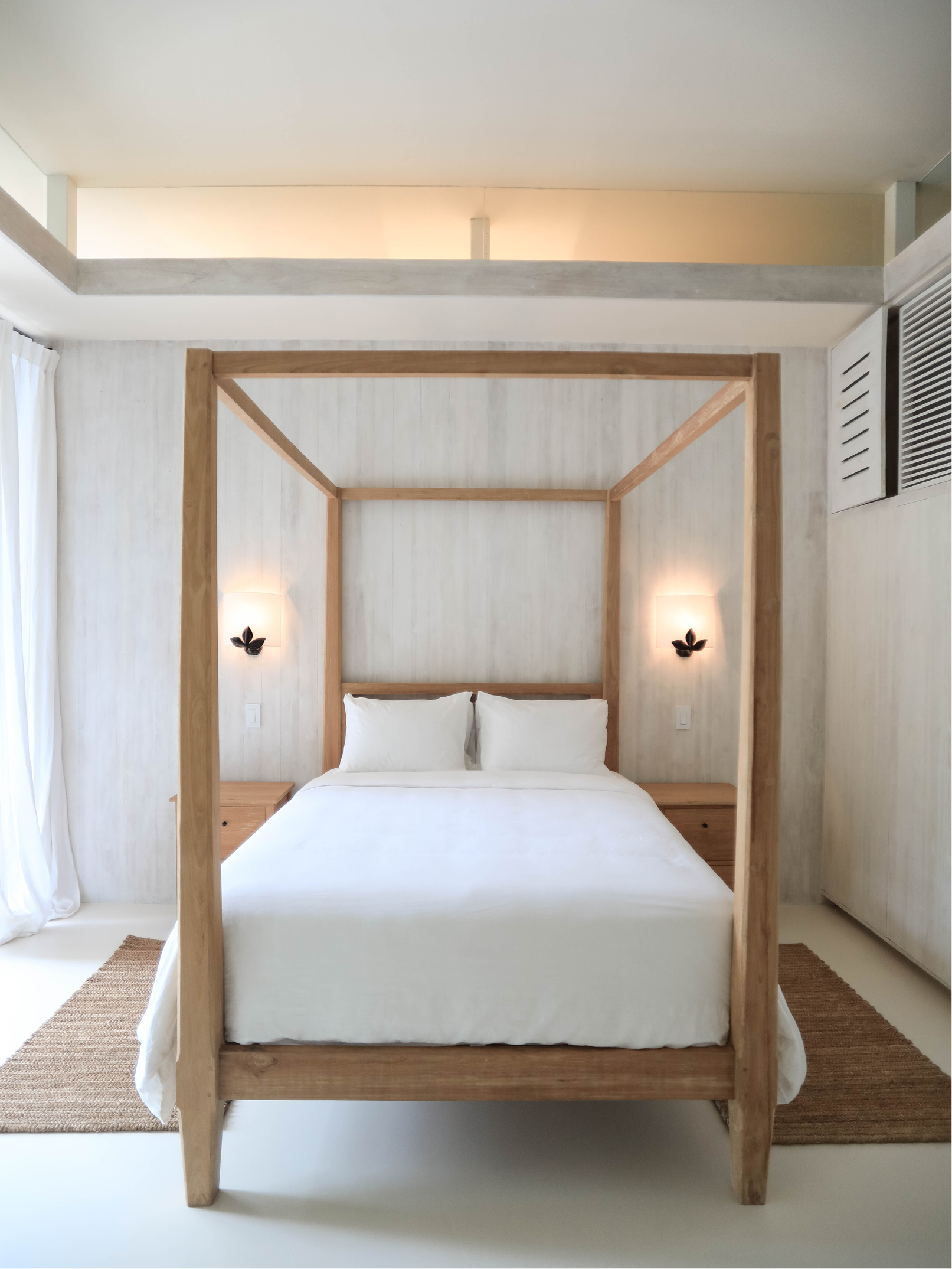 A bedroom with a white bed, wooden canopy frame, beige area rug, and wall-mounted black lamps with floral designs.