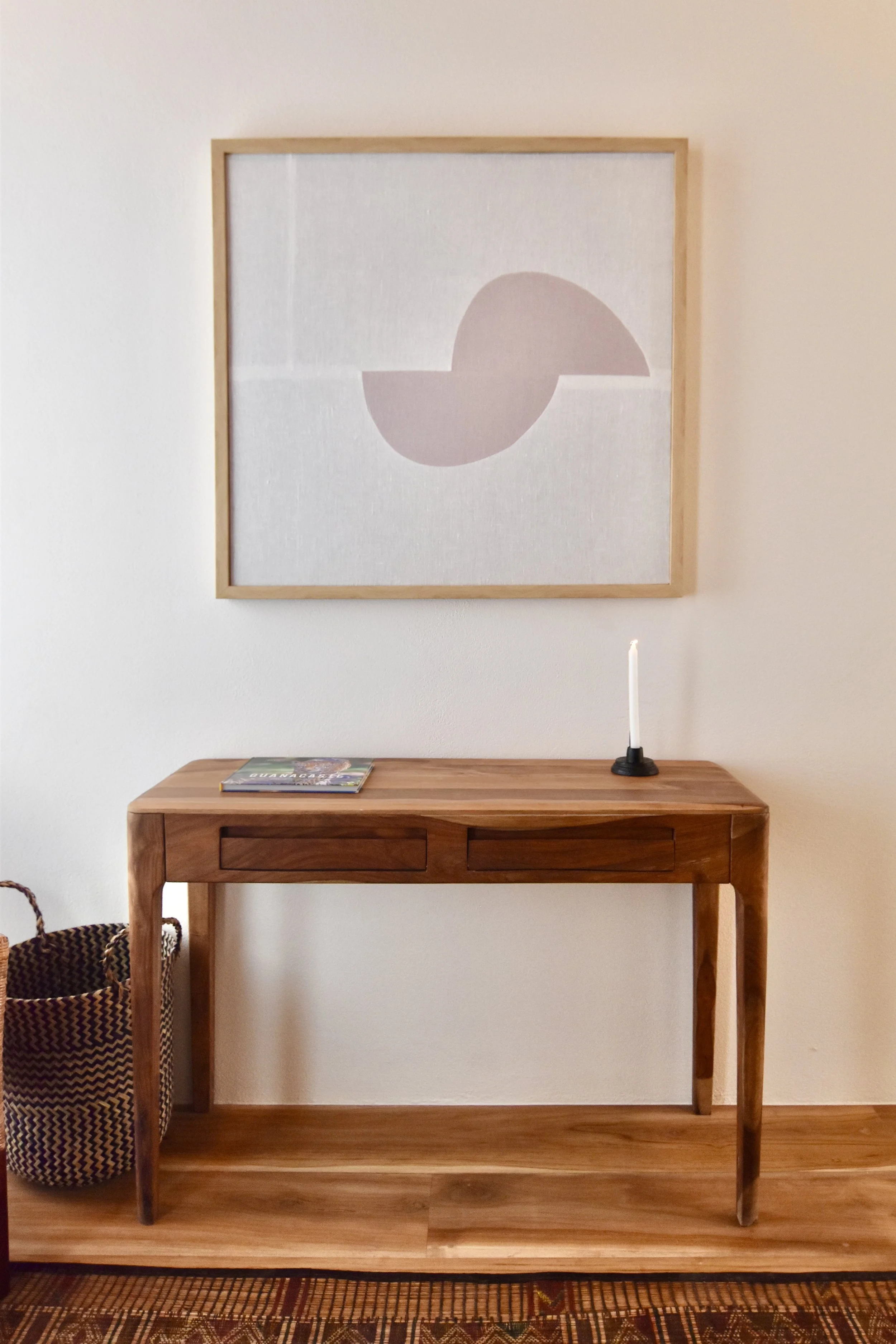 Wooden console table with a candle and magazine, a framed abstract artwork with geometric shapes, and a woven basket beneath, against white walls and hardwood floors.