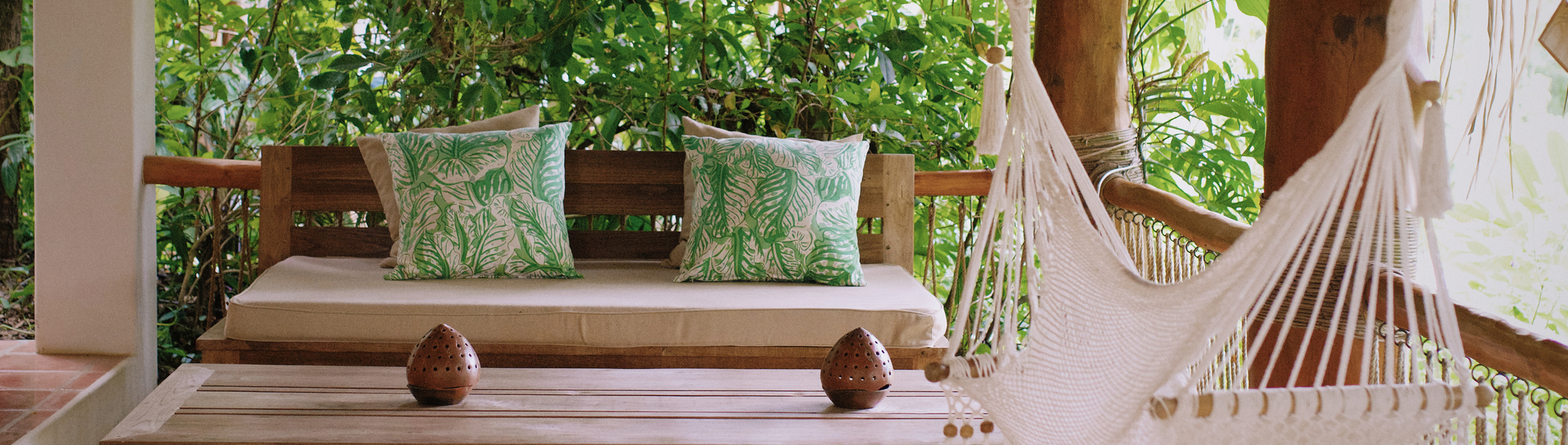 Outdoor patio area with a wooden bench with green and white leaf-patterned cushions, a white hammock chair, and two small decorative objects on a wooden table, surrounded by lush green foliage.