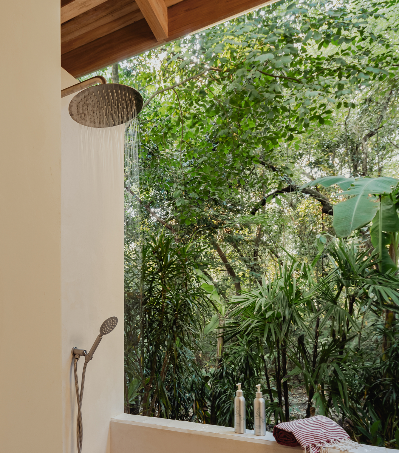 Outdoor shower area with a showerhead and a hand-held shower fixture mounted on a light-colored wall, overlooking lush green trees and plants, with two bottles and a folded towel on the ledge.