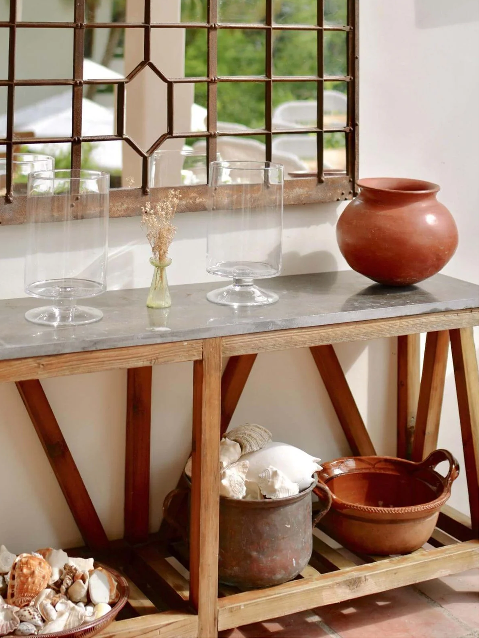 A wooden shelf holding glass containers, seashells, and large clay pots with an open window showing greenery outside.
