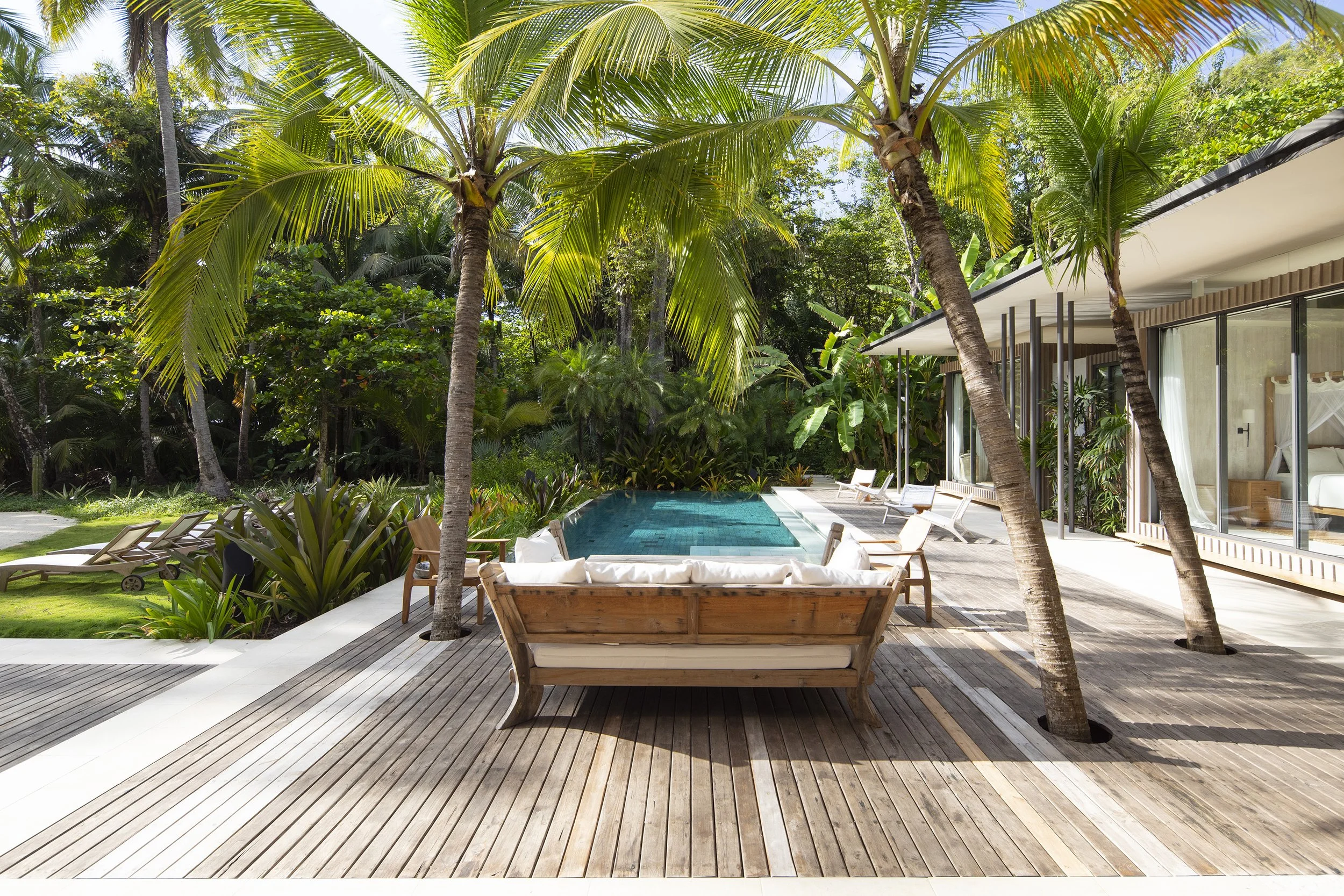 Luxury modern house with floor-to-ceiling glass windows overlooking a tropical backyard with tall palm trees, a swimming pool, and outdoor lounge chairs on a wooden deck.