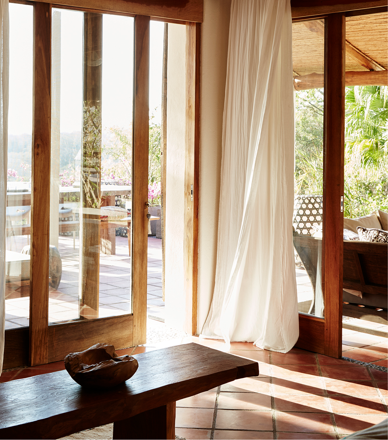 Open glass sliding doors leading to an outdoor patio with wooden furniture and trees in the background. Inside, a curtain hangs next to the doors, and a wooden table with a decorative bowl is in the foreground. Sunlight streams into the room.