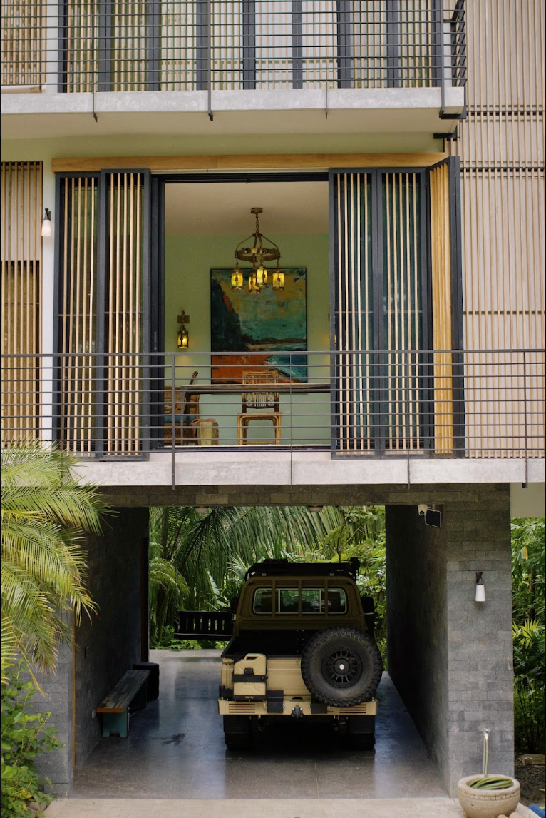 A modern house with an open garage underneath, containing a beige off-road vehicle, and a second floor with a balcony featuring vertical wooden slats and a view of a green wall with artwork and hanging lights inside.