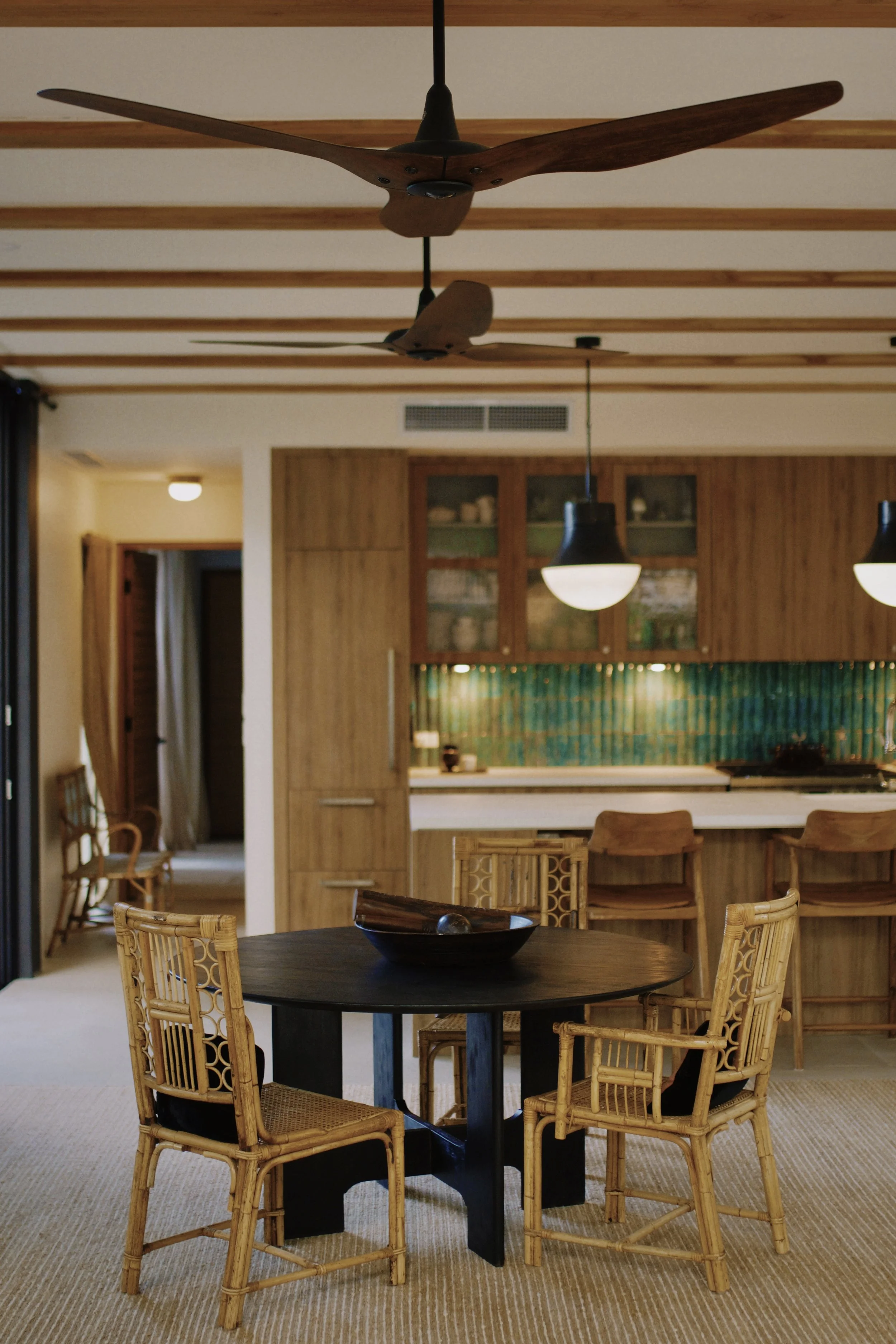 A dining room with round black table and six wooden chairs, a kitchen area in the background with cabinetry and pendant lighting, and a ceiling fan with wooden blades.