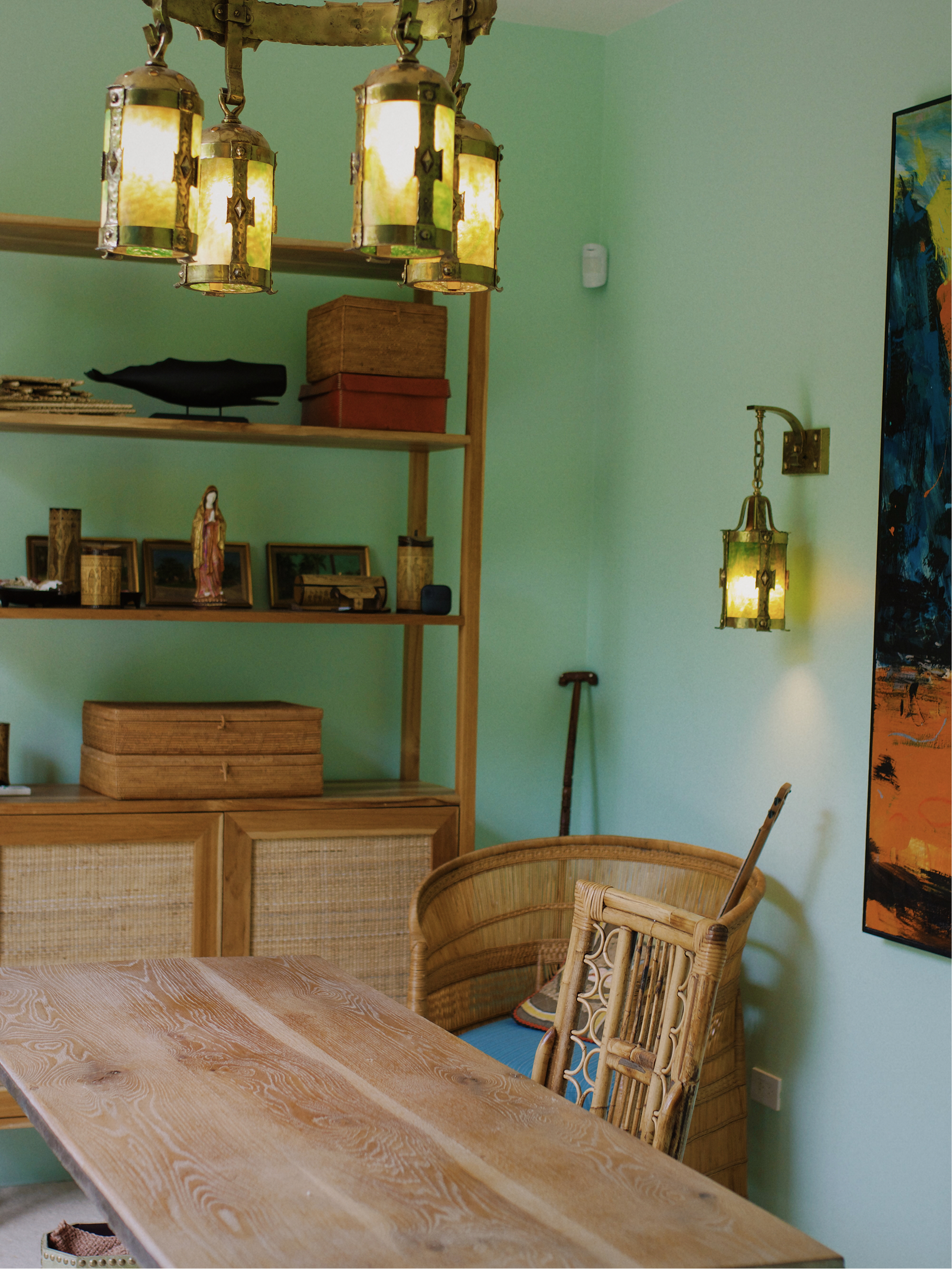 A dining room with a wooden table, woven chairs, a bookshelf with folk art, and decorative hanging lamps with stained glass panels.