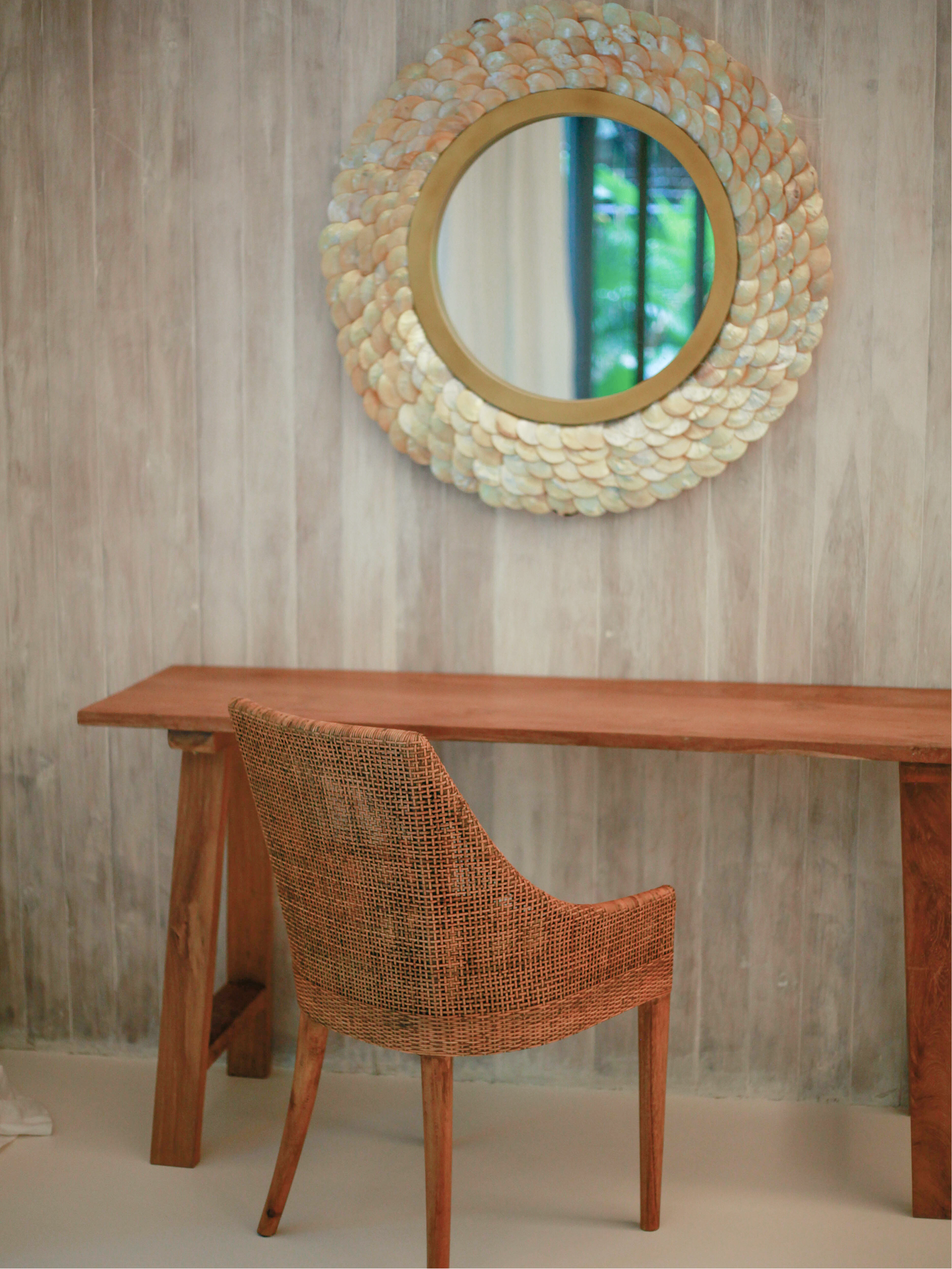 A rattan chair with a curved backrest in front of a wooden table, behind a round wall mirror with a shell frame, against a textured beige wall.