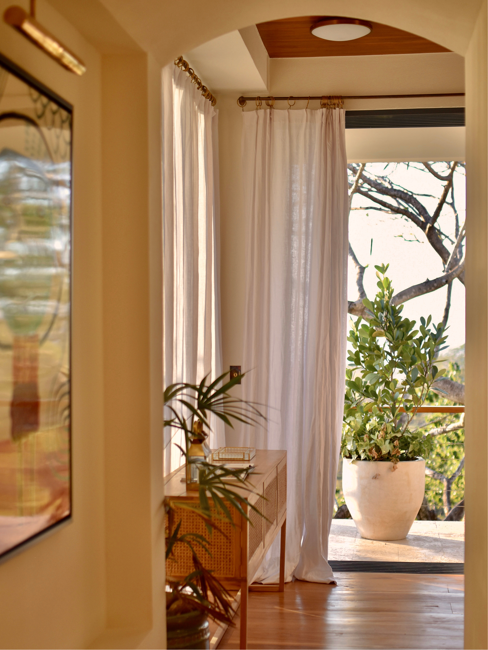 A cozy indoor-outdoor space with large potted plant, wooden furniture, white curtains, and sunlight coming through a window.