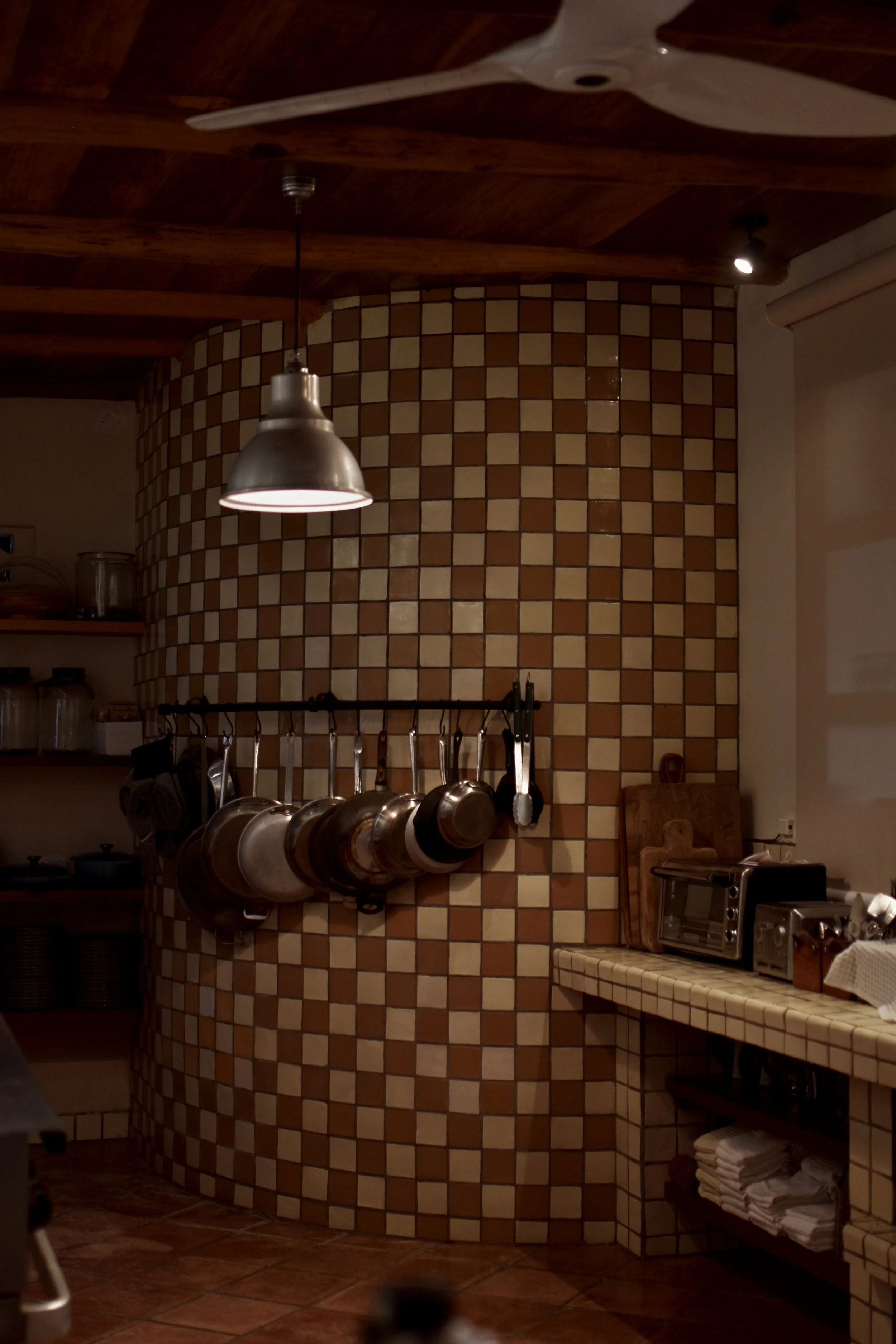 A kitchen with a tiled curved wall, hanging pots and pans, a wooden countertop with a toaster oven, and shelves with dishes and kitchen utensils.