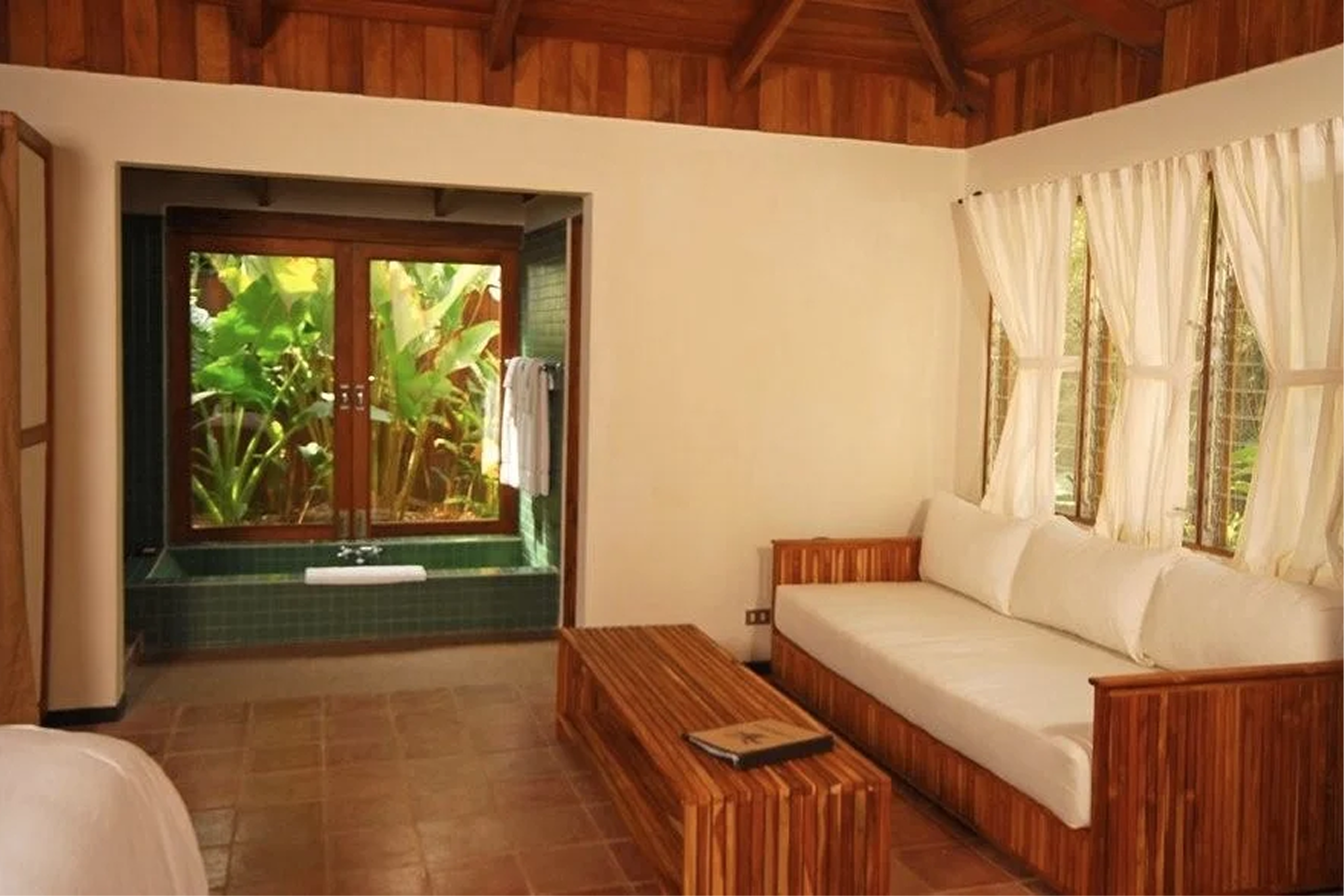 A living room with wooden ceiling beams, beige walls, a large window with white curtains, and a sliding glass door leading outside with lush green plants visible. There is a white sofa with wooden accents and a wooden table with a magazine on it.