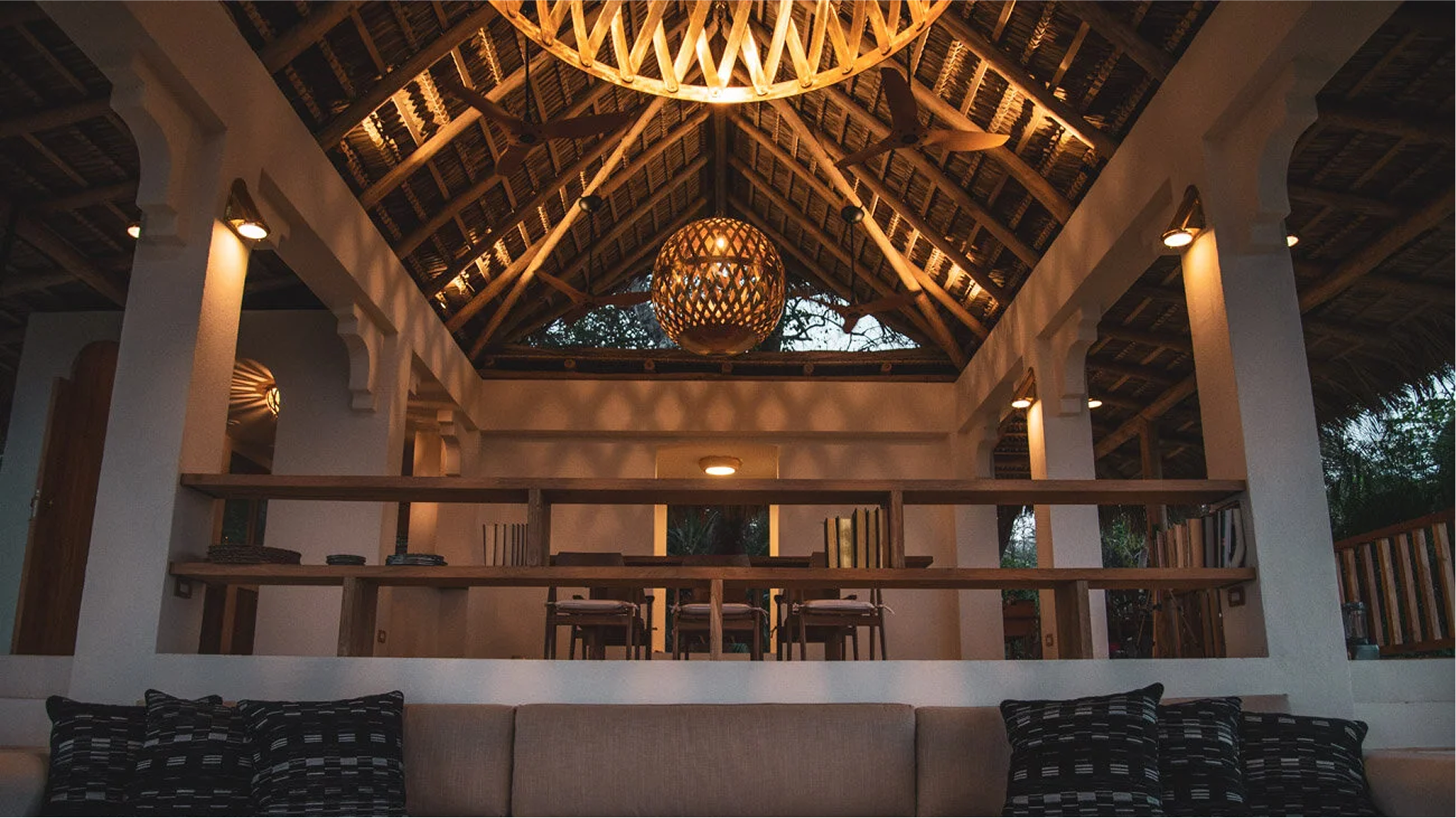 Interior view of a building with a high, wooden, thatched roof supported by white columns. There are hanging circular woven light fixtures and ceiling fans. The space appears open and airy with natural light coming through the open ceiling area.