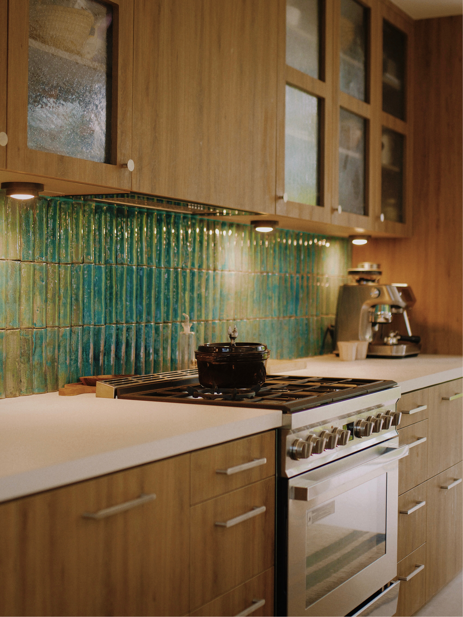 A kitchen with wooden cabinets, a green tiled backsplash, a stove with a black pot, and a coffee machine on the counter.