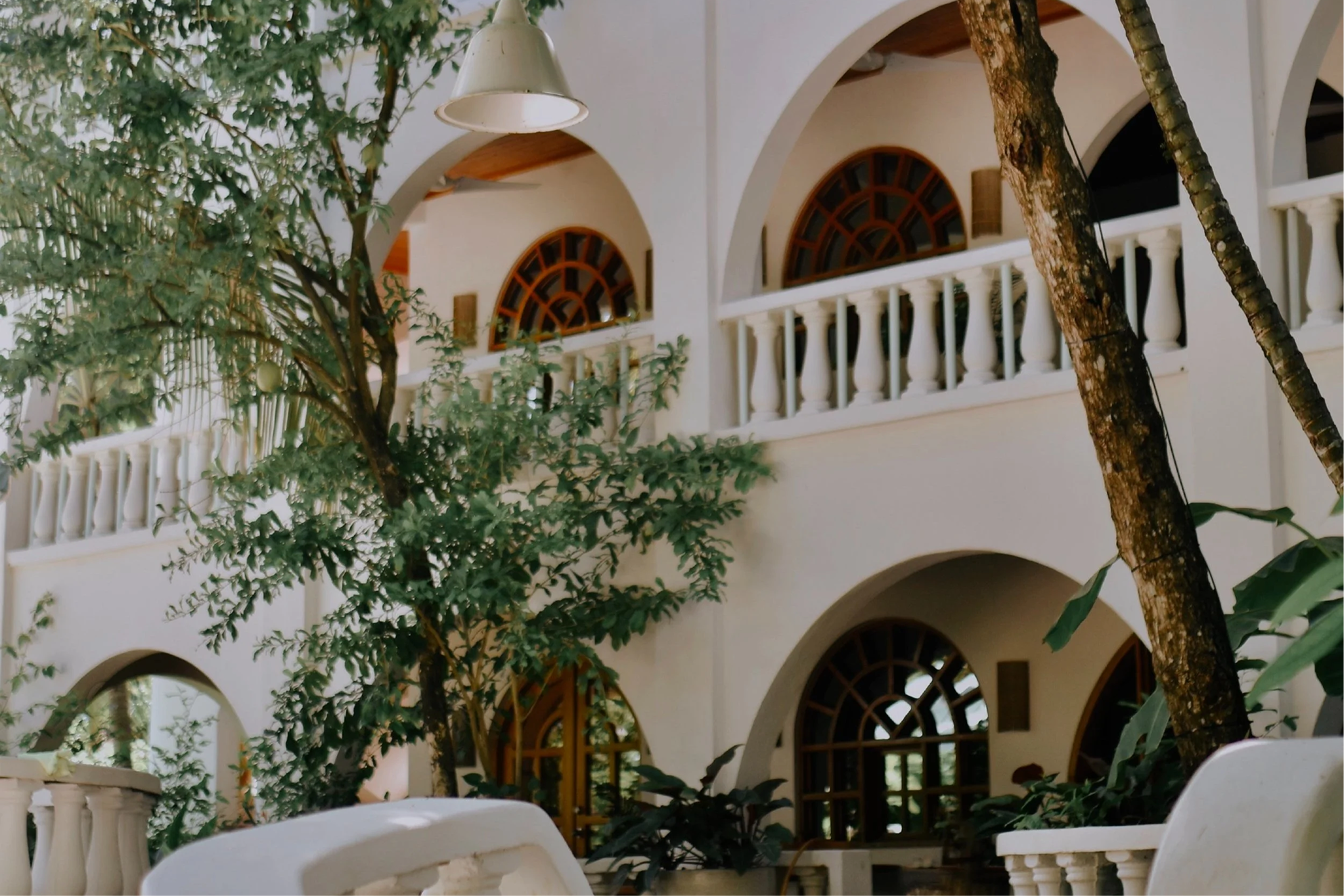 Exterior view of a Mediterranean style house with white walls, arched windows, and a white balcony railing, partially obscured by green trees.