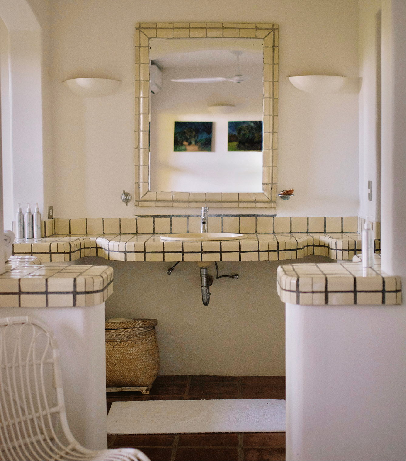 Bathroom with beige tiled vanity, mirror, and white walls. Contains bottles and a wicker basket underneath, mirror reflects art paintings on the opposite wall.