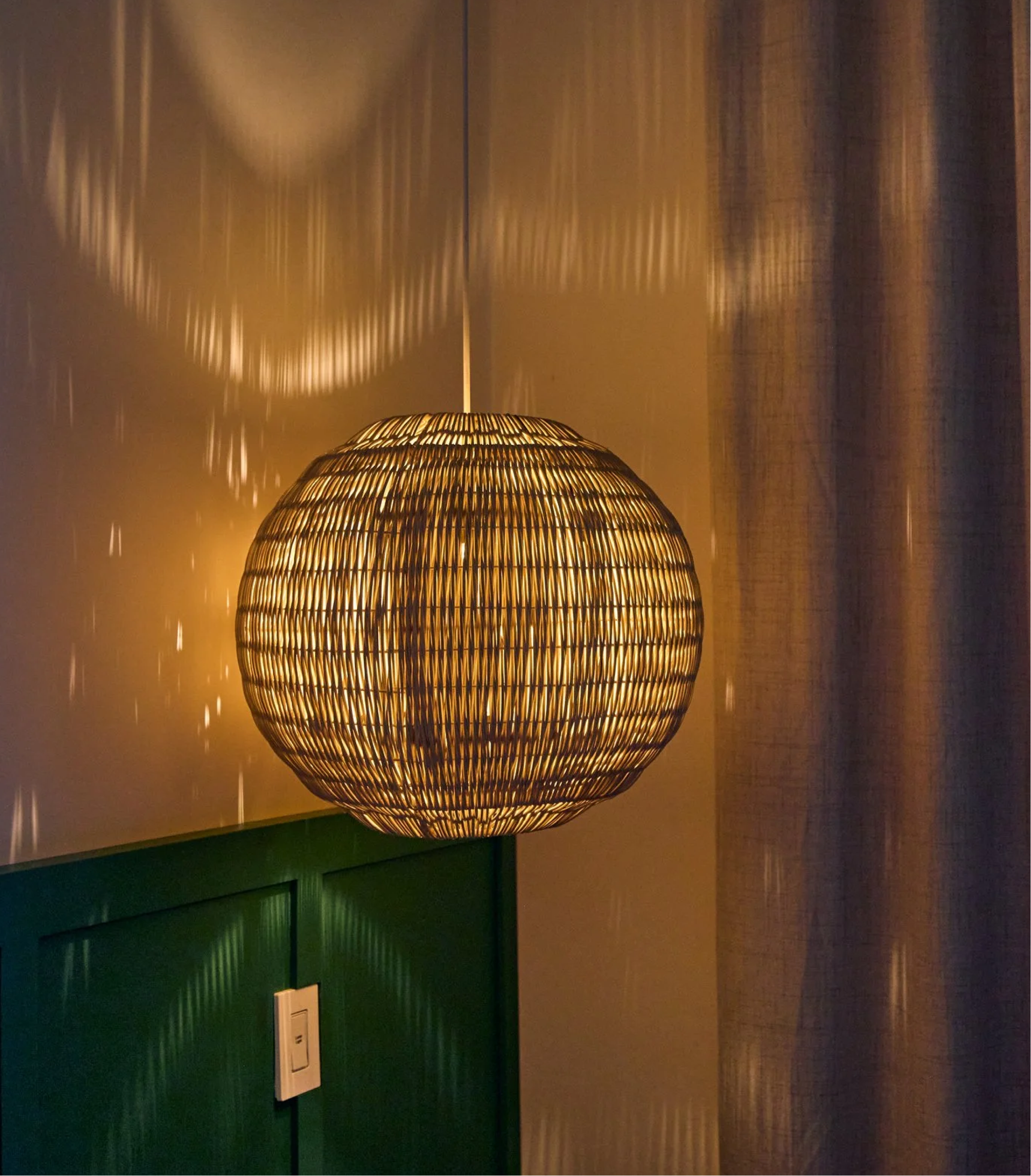 A ceiling lamp with a woven bamboo shade casting warm light and shadows on the ceiling, mounted above a dark green cabinet in a room with wooden flooring.