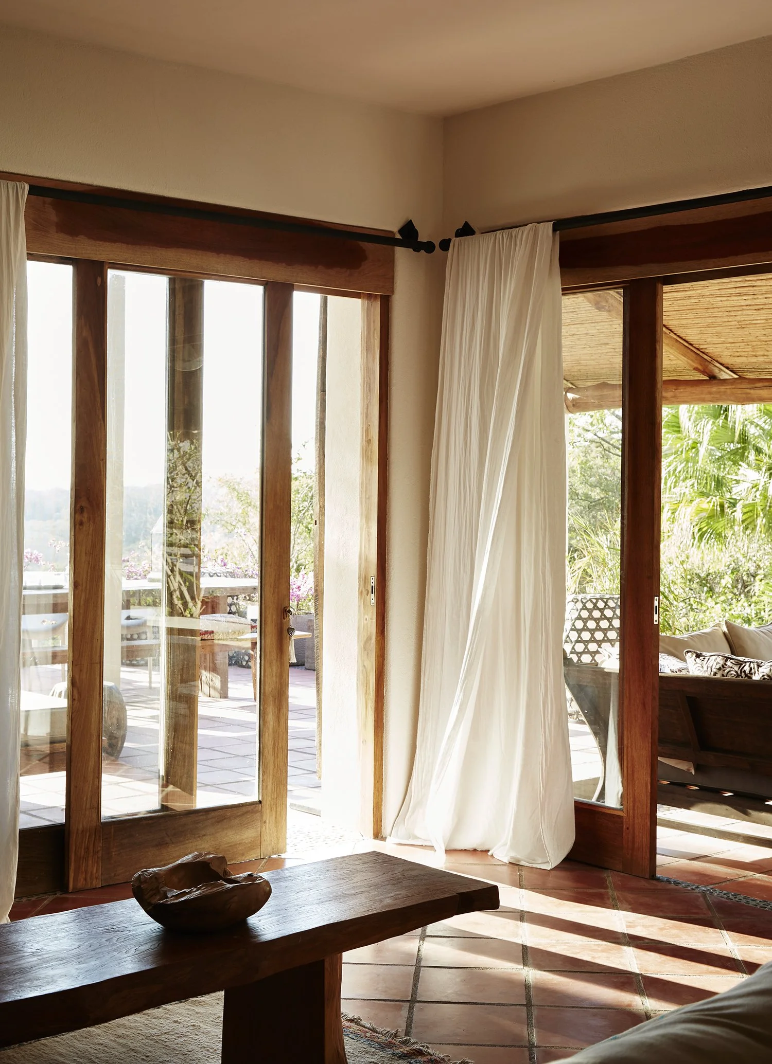 Sunlit living room with large glass sliding doors, white curtains, wooden frames, and outdoor patio with plants visible outside.