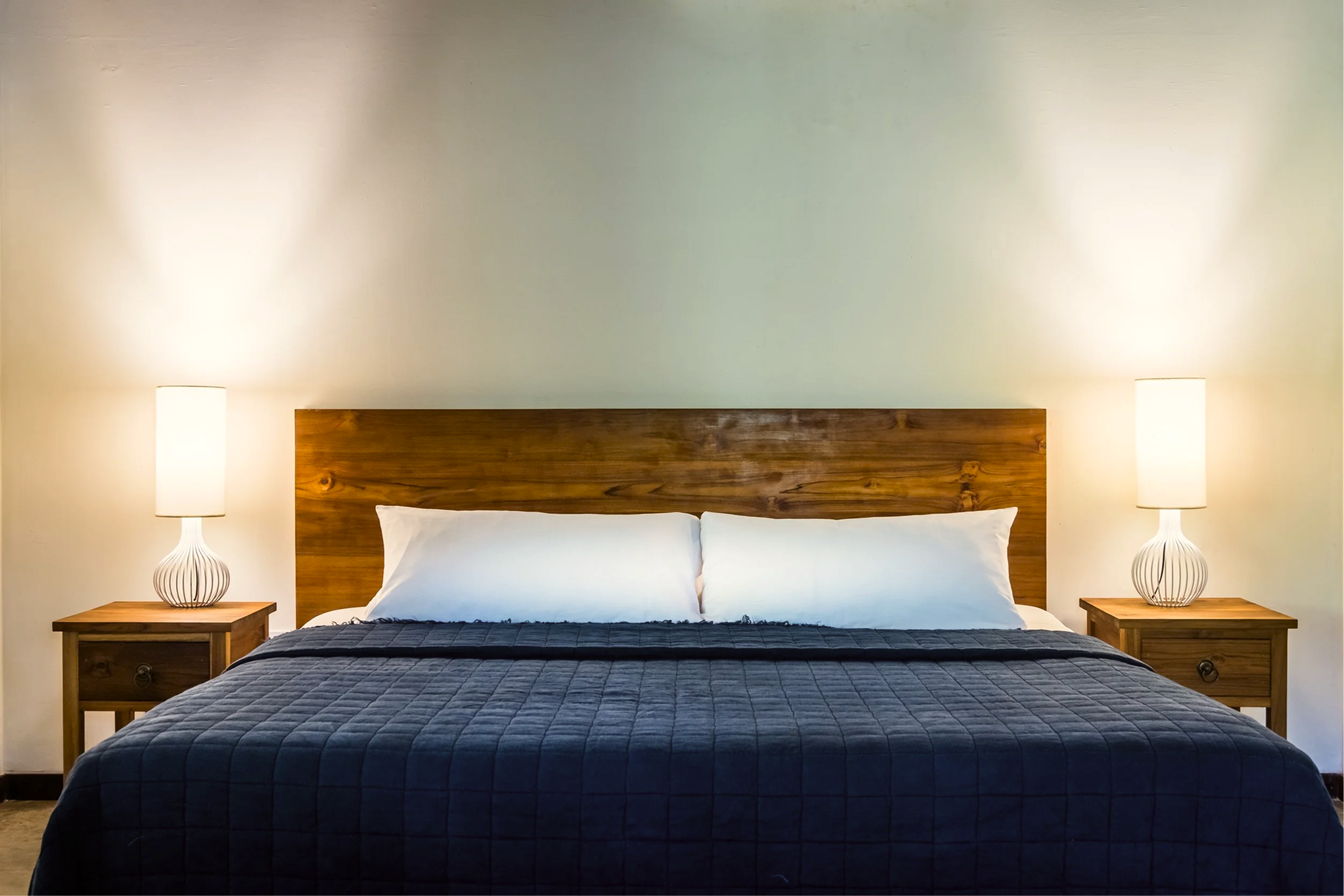 A neatly made bed with white pillows and a dark quilt, a wooden headboard, and matching nightstands with white lamps on each side.