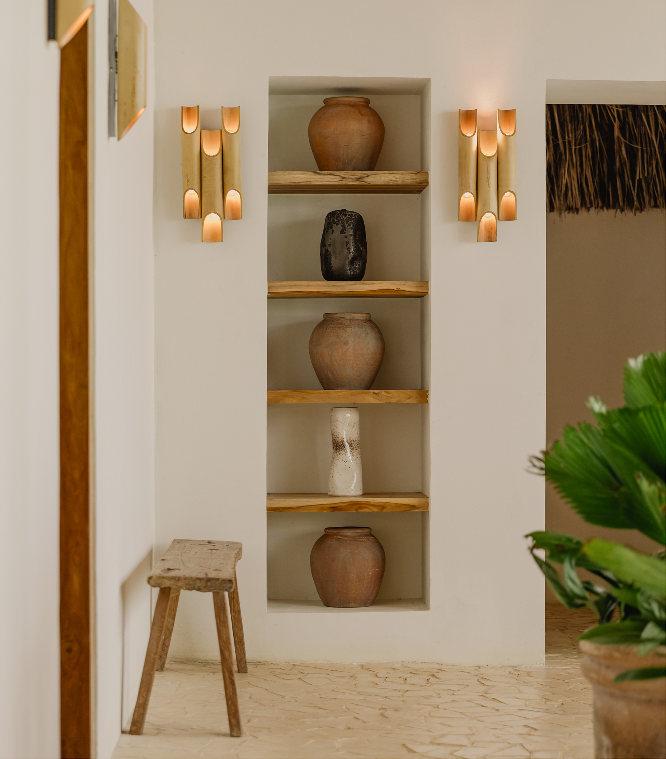 Interior of a room with a built-in shelf containing three clay vases, a rustic wooden stool, and wall-mounted bamboo light fixtures on either side of the shelf. Part of a plant is visible in the foreground.