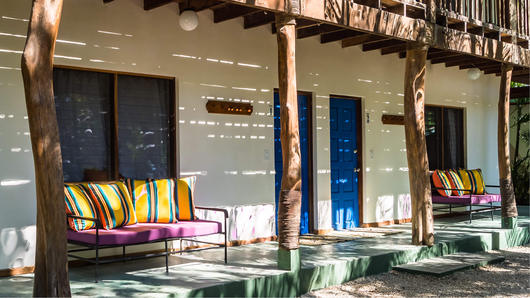 Porch area outside a building with blue doors, timber columns, colorful striped cushions on pink seating, and sunlight creating shadows on the wall.