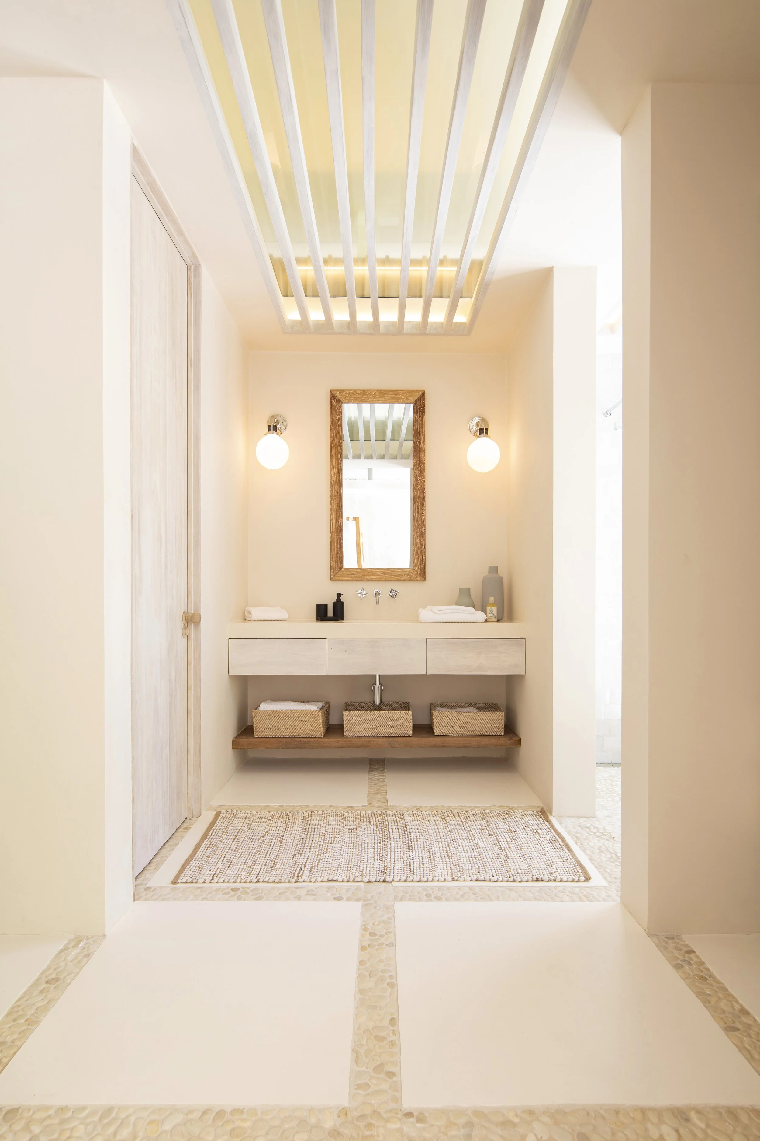 A minimalist bathroom with a white floating vanity, a mirror with a wooden frame, two wall-mounted light fixtures, and decorative baskets on a wooden shelf. The room has a soft, neutral color palette with beige walls, a textured rug, and a ceiling wi