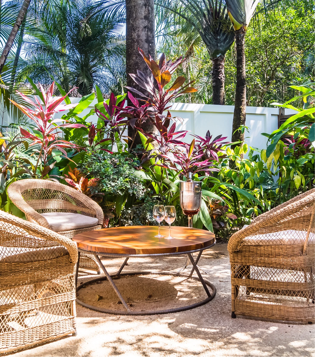 Outdoor patio with wicker chairs around a wooden table, two wine glasses, a metal ice bucket, lush tropical plants, tall trees, and a white fence in the background.