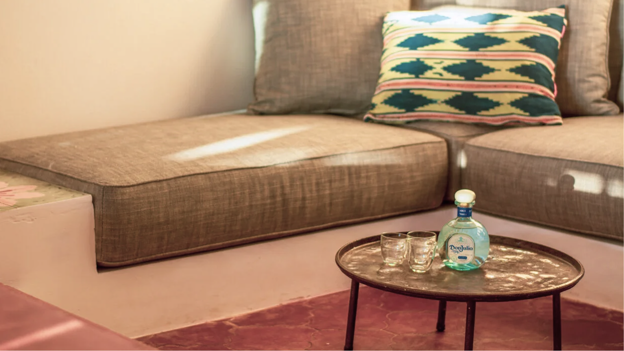 A living room with a brown sofa, three cushions, and a small round table holding a bottle of Don Julio tequila, two shot glasses, and a glass. The room is illuminated with natural light.