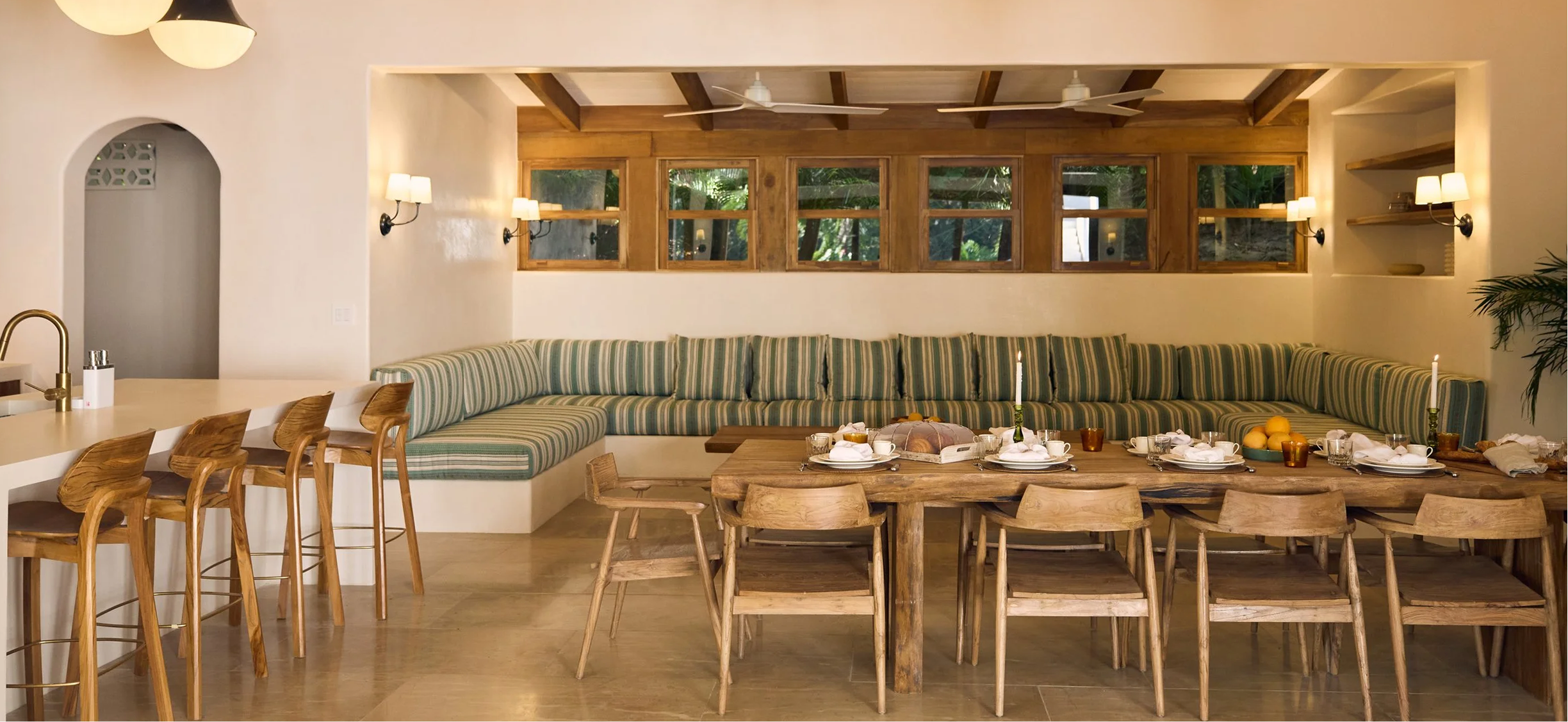 A cozy dining area with a large wooden table set for a meal, surrounded by wooden chairs, with a built-in curved bench with striped cushions along the wall. Candles, a loaf of bread, fruit, and tableware are arranged on the table. The room has a warm, tropical style with wooden accents, wall sconces, and a view of greenery through multiple small windows.