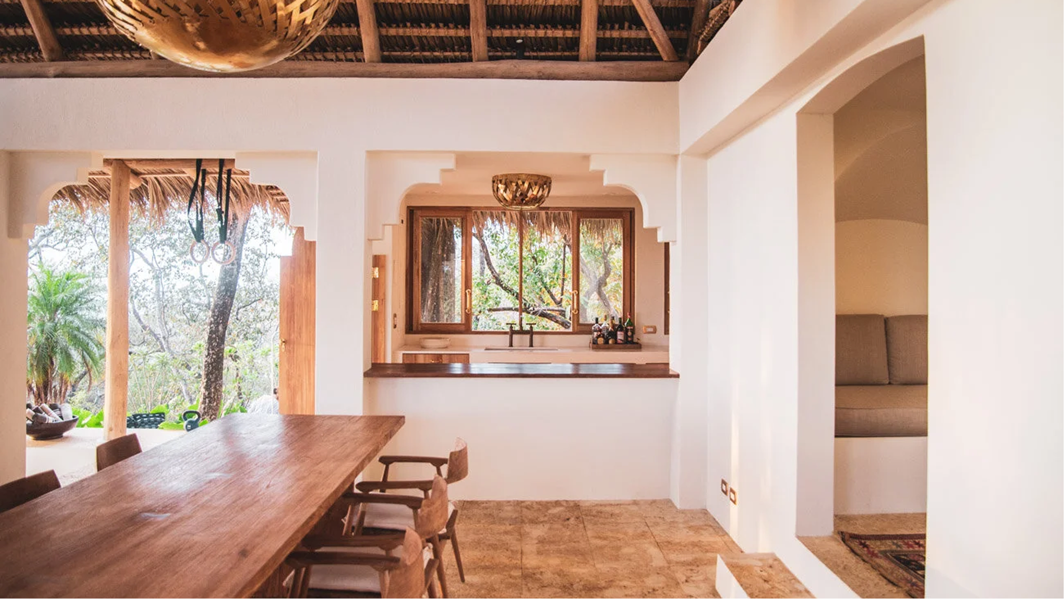 A cozy interior of a house with a wooden dining table and chairs, open kitchen with a window overlooking trees, and natural sunlight streaming inside.