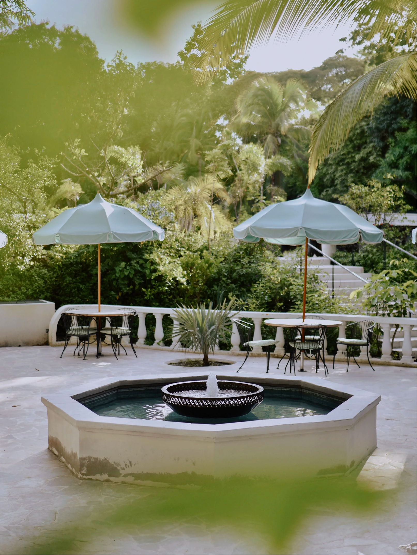 Outdoor patio with white umbrellas, black tables and chairs, a fountain in the foreground, surrounded by lush green trees and plants.