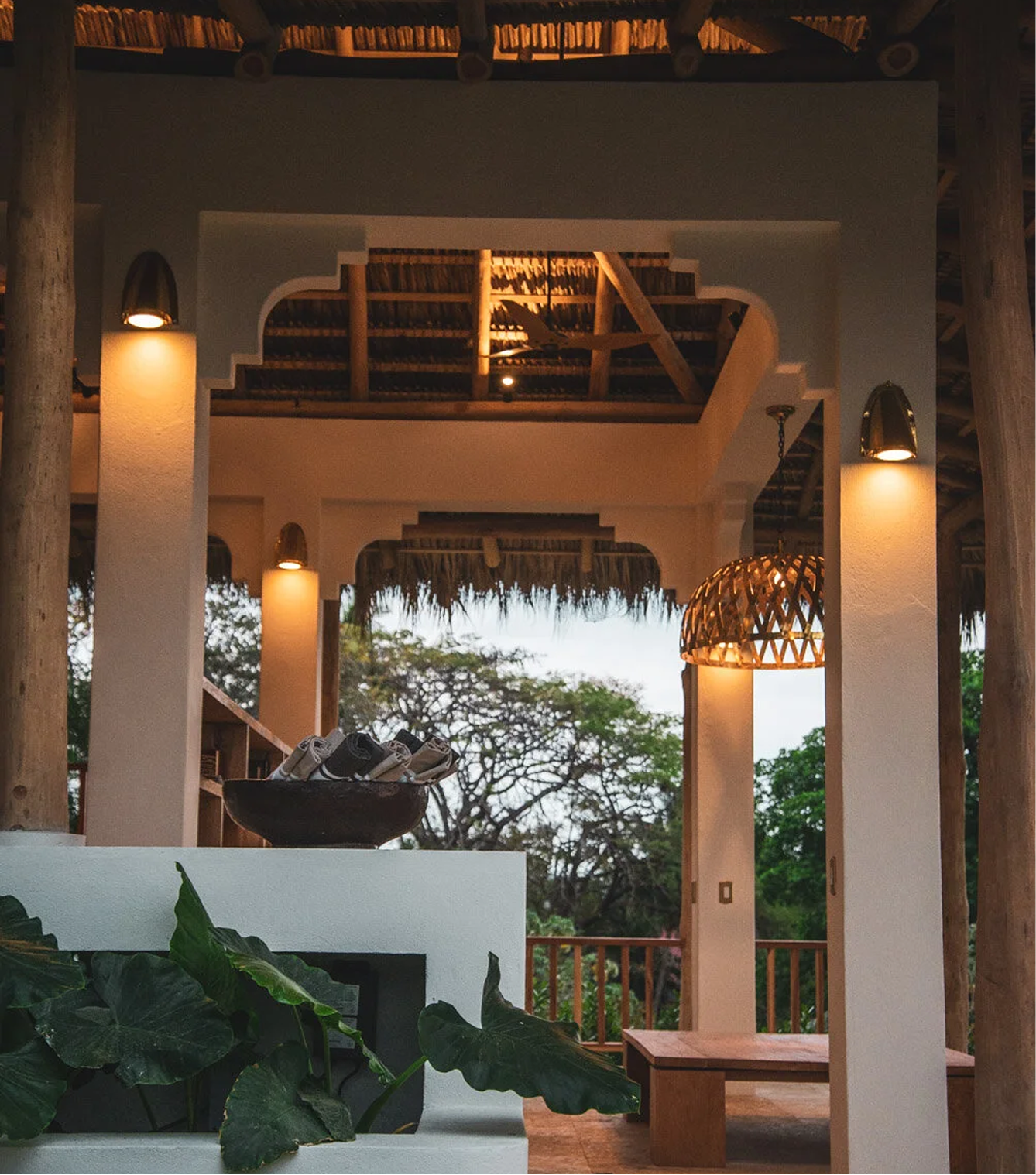 Interior of a rustic open-air space with wooden beams, thatched roof, decorative lighting, and lush greenery outside.