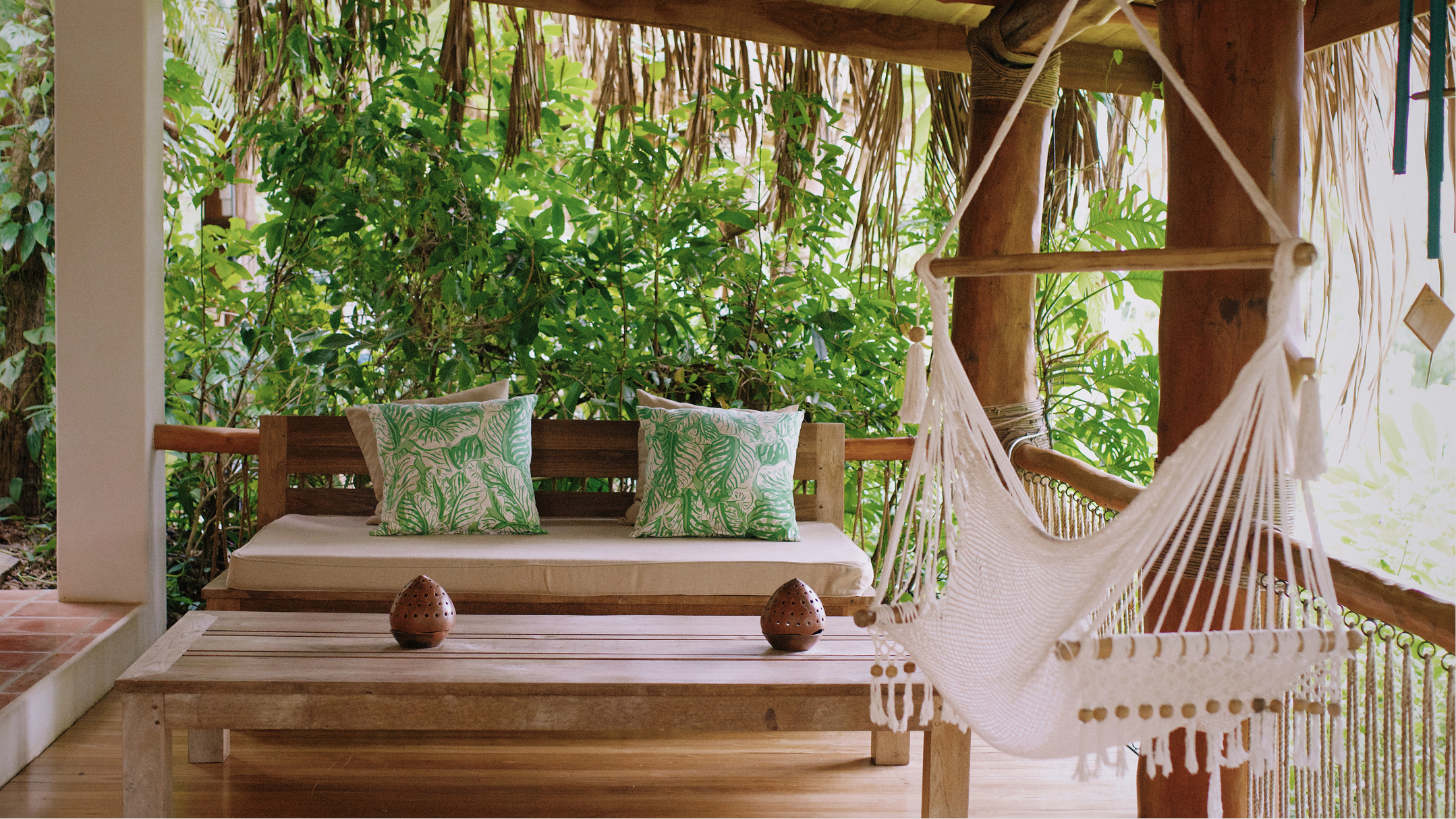 A wooden porch or deck area with a cushioned bench, green tropical-patterned pillows, a hammock chair, and lush green foliage in the background.