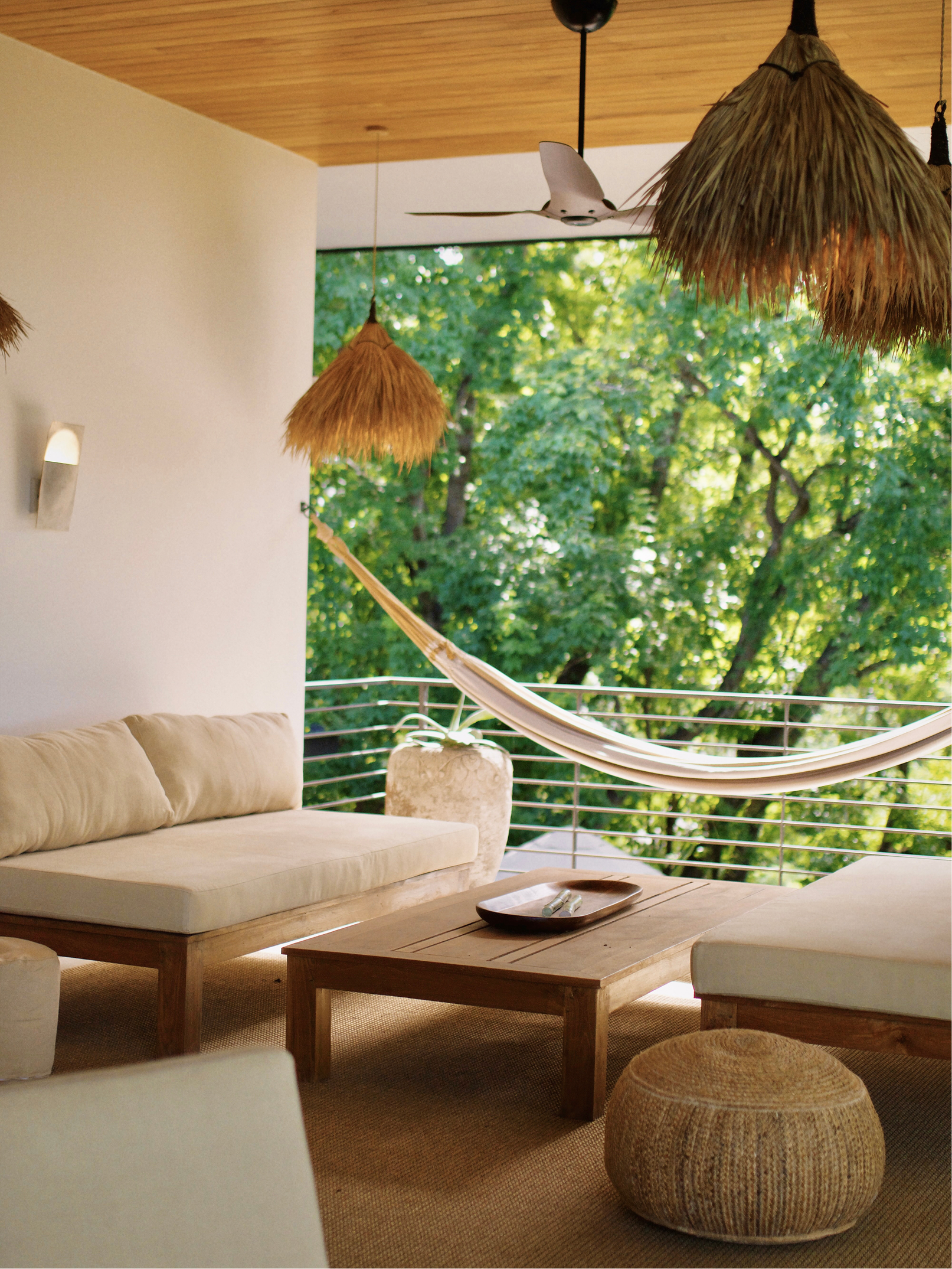 Relaxing outdoor living space with a beige sofa, wooden coffee table, wicker ottoman, and hammock, surrounded by lush green trees, under a wooden ceiling with hanging straw pendant lights and a ceiling fan.