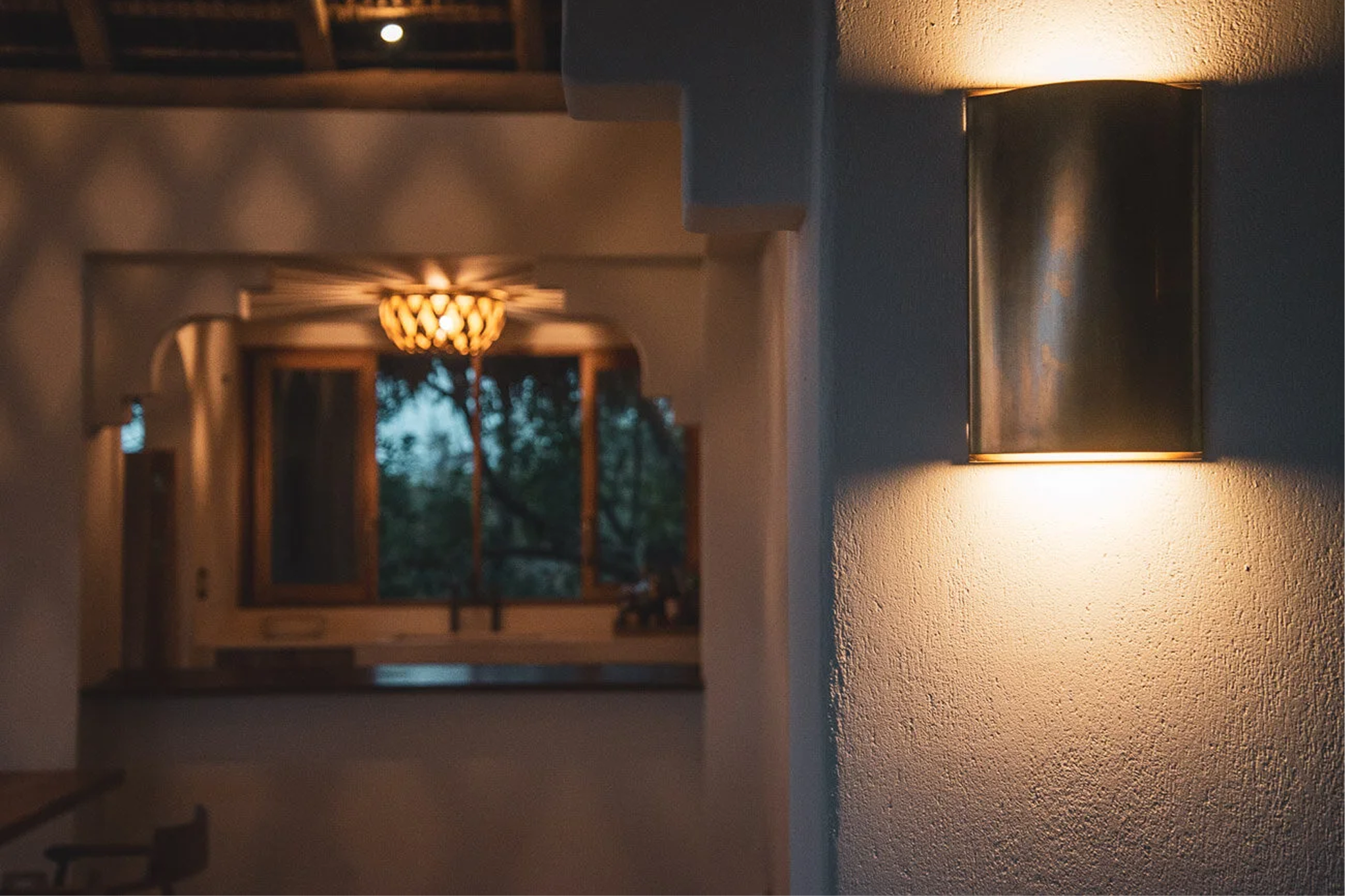 Interior view of a home with a modern wall-mounted light fixture and a hallway that leads to a room with a window, trees outside, and a hanging ceiling light.