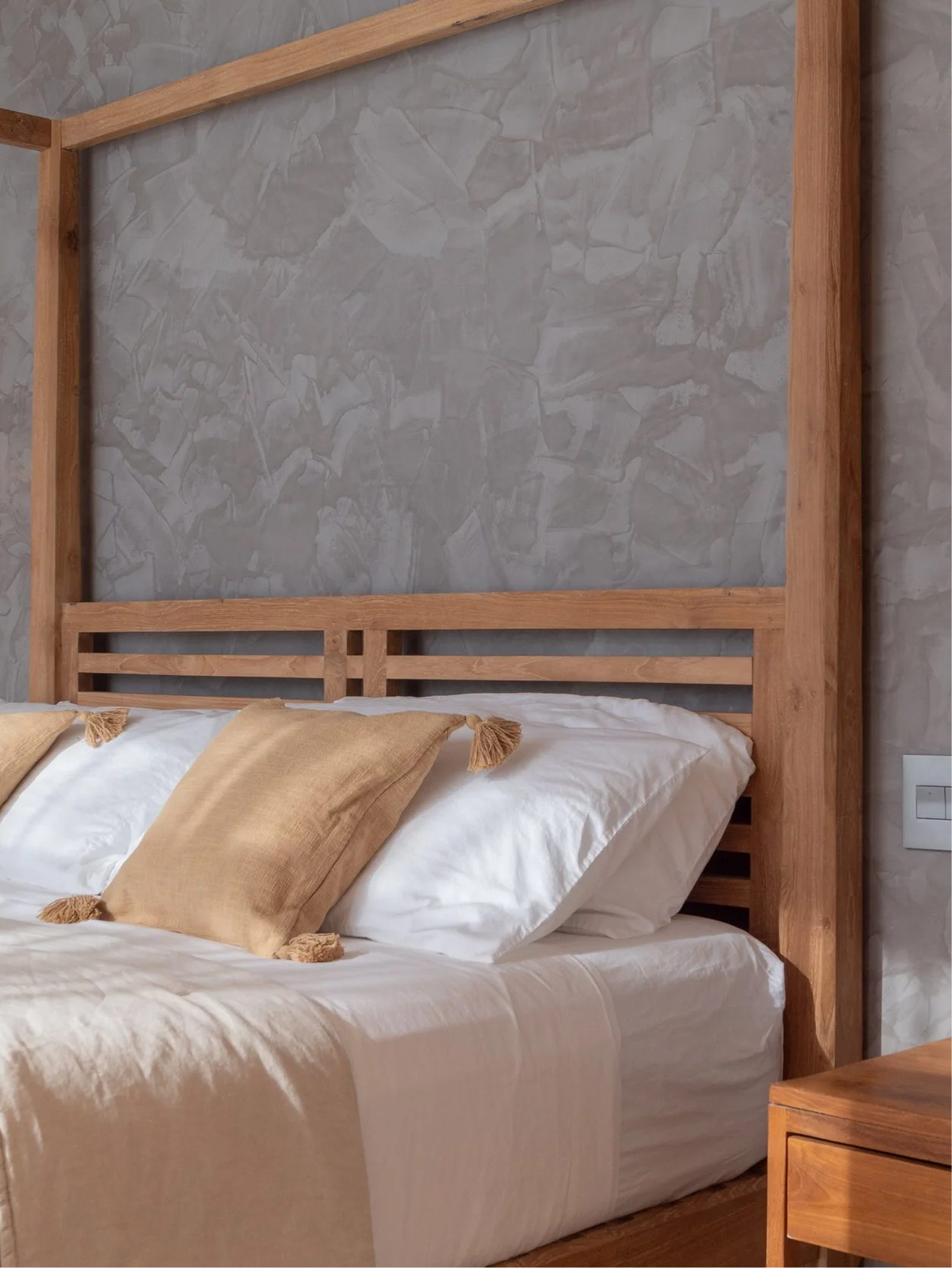 A wooden bed frame with a white mattress and beige pillows with tassels, set against a gray textured wall.