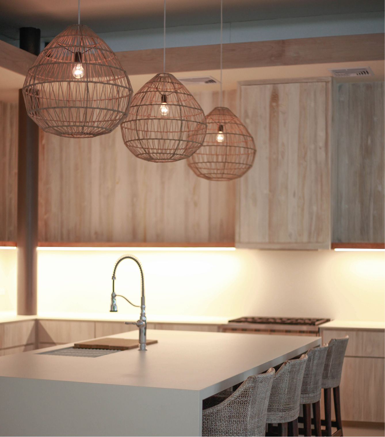 Modern kitchen with three wicker pendant lights hanging above a white island with a sink. Wooden cabinets and a marble top stove are visible.