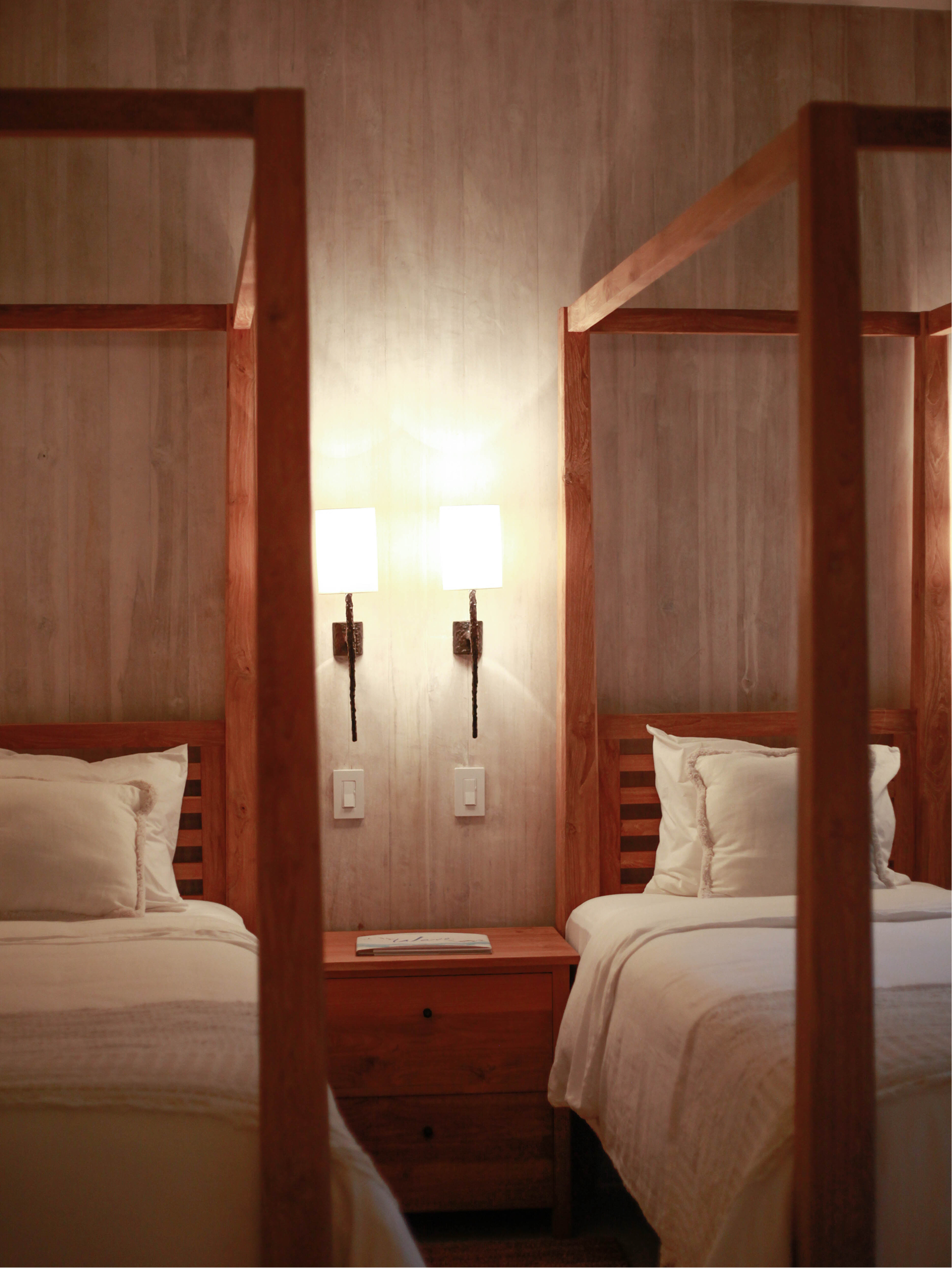 Two beds with white linens and pillows in a hotel room, separated by a wooden nightstand, with two lamps on the wall behind them.