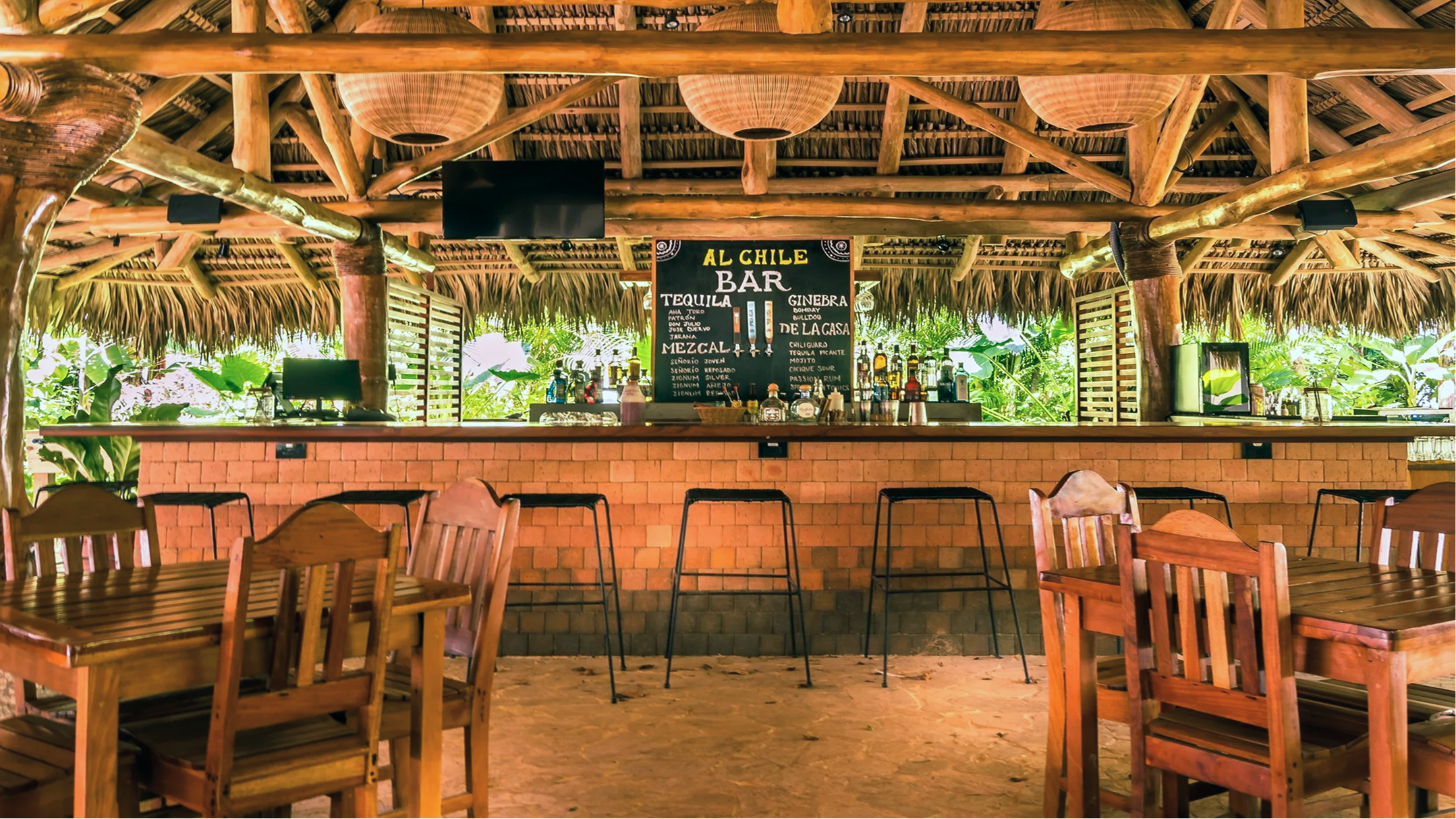 Bar area with a thatched roof, wooden beams, and a chalkboard menu listing drinks like tequila, gin, and mezcal. There are wooden tables and chairs in the foreground, lush greenery visible behind the bar, and several bottles and glasses on the bar counter.