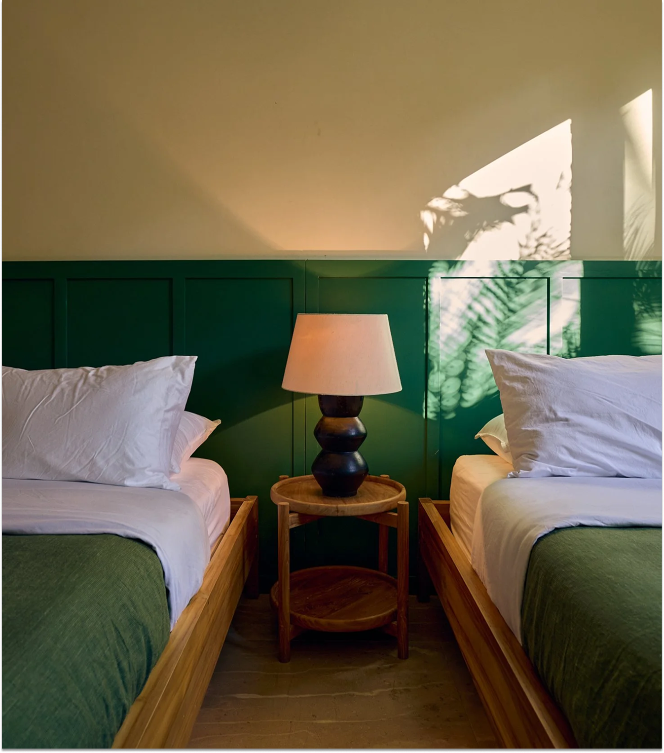 Two beds with white pillows and green bedding, a wooden nightstand in between with a black lamp, against a green paneled wall. Shadows of leaves are projected onto the wall.