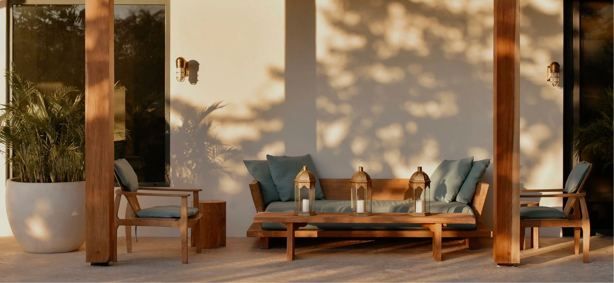 A cozy outdoor seating area with a wooden couch and chairs with cushions, a wooden table with three lanterns, potted plants, and sunlight casting patterns on the wall.