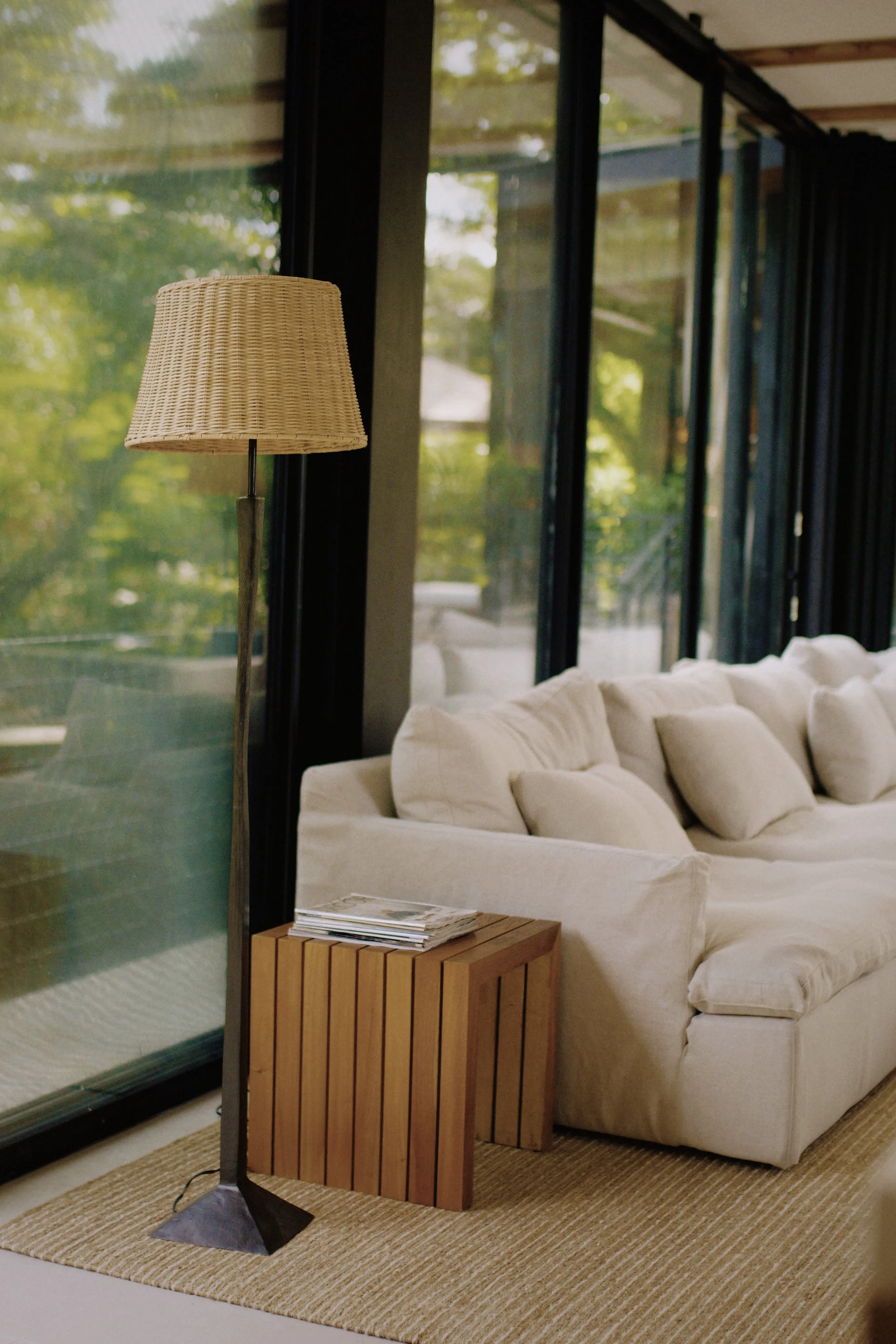 Living room with white sofa, side table with magazines, floor lamp, large windows, and green trees outside.