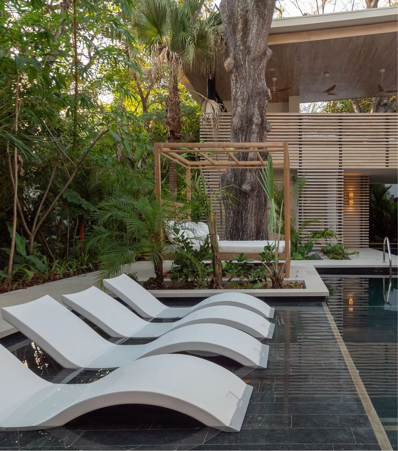 Modern outdoor pool area with white lounge chairs, lush green plants, a large tree, and a wooden swing, with a building featuring horizontal wooden slats in the background.